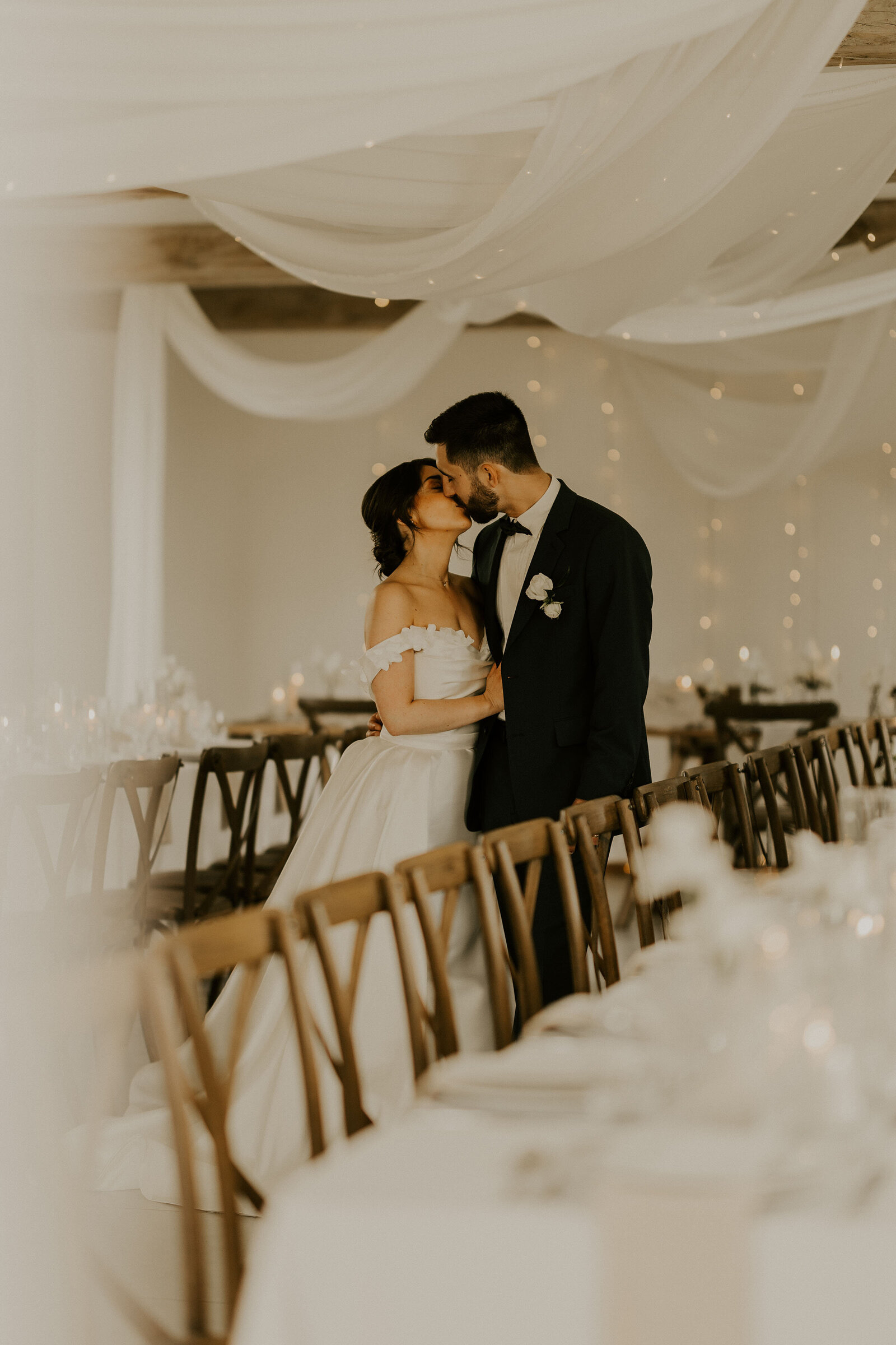 Mariés échangeant un baiser dans leur salle de repas décorée de chaises en bois, de lumières et de voilage blanc en attendant les invités. Photographie de mariage réalisée par Laura Termeau photographie.