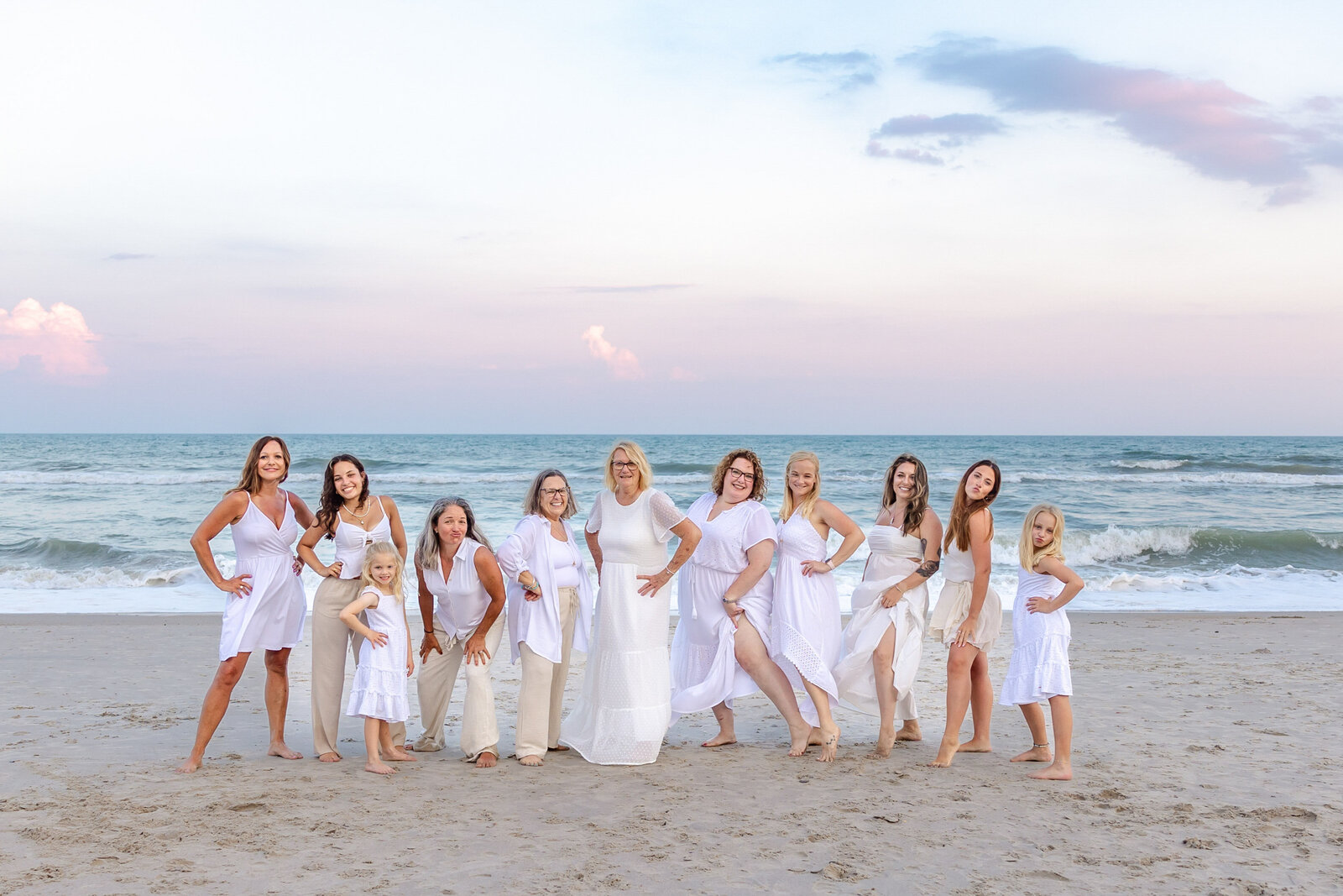 A group of women and girls dressed in elegant white outfits pose gracefully on Wrightsville Beach. The soft pastel sky and ocean waves create a serene and timeless backdrop. Ideal for destination family photographer NC, capturing elegant beachside moments.