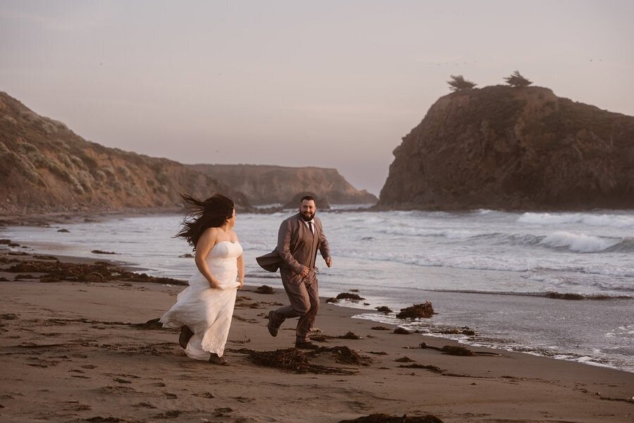 A bride and groom run along the shoreline at Big Sur, laughing as the waves crash behind them and the wind blows through the bride’s hair.