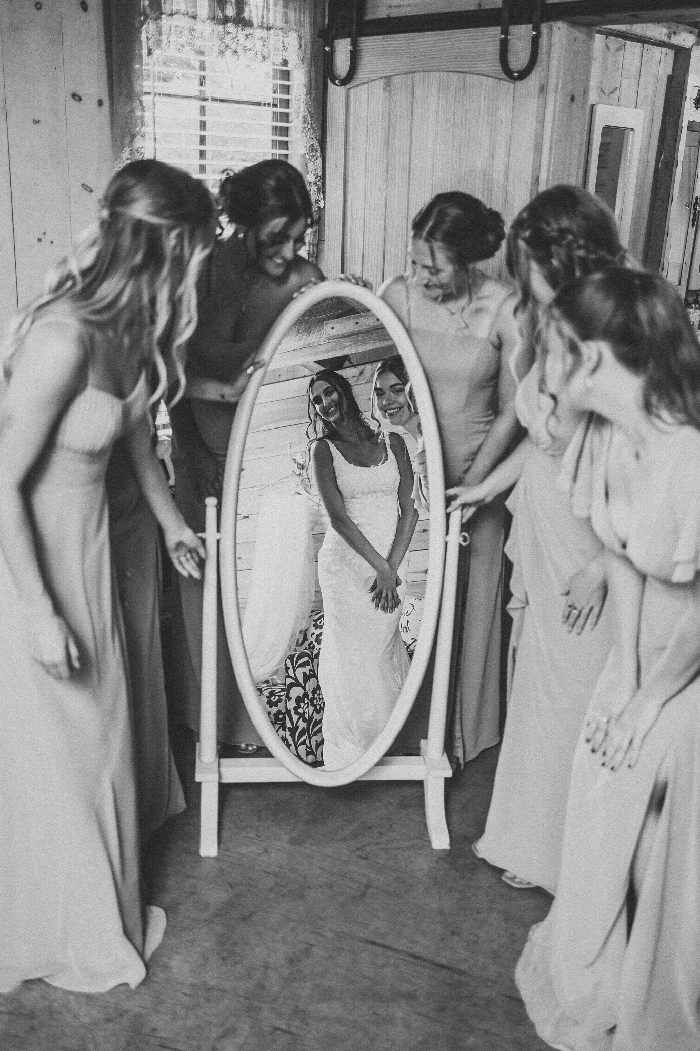 A bride looks at her reflection in a full length mirror surrounded by her bridesmaids lookin in awe of her beauty