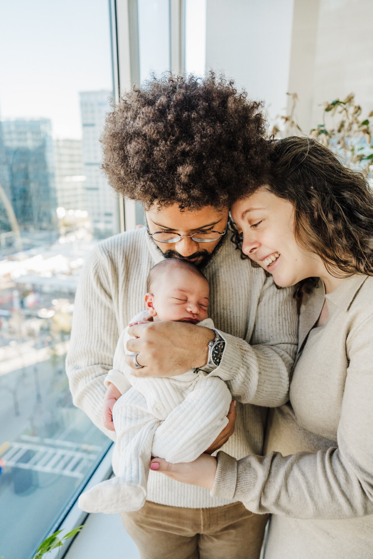 new parents dressed in beige stand by window of a high rise apartment with their new baby