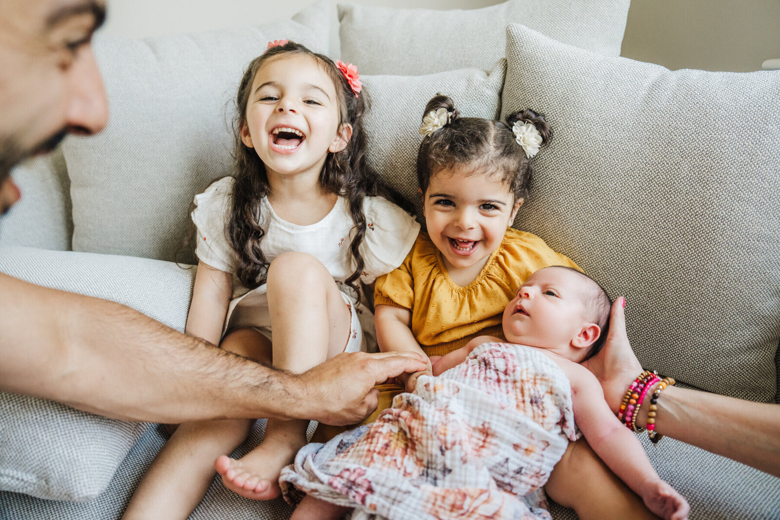 two big sisters laugh as mom and dad hand newborn sister to them on couch