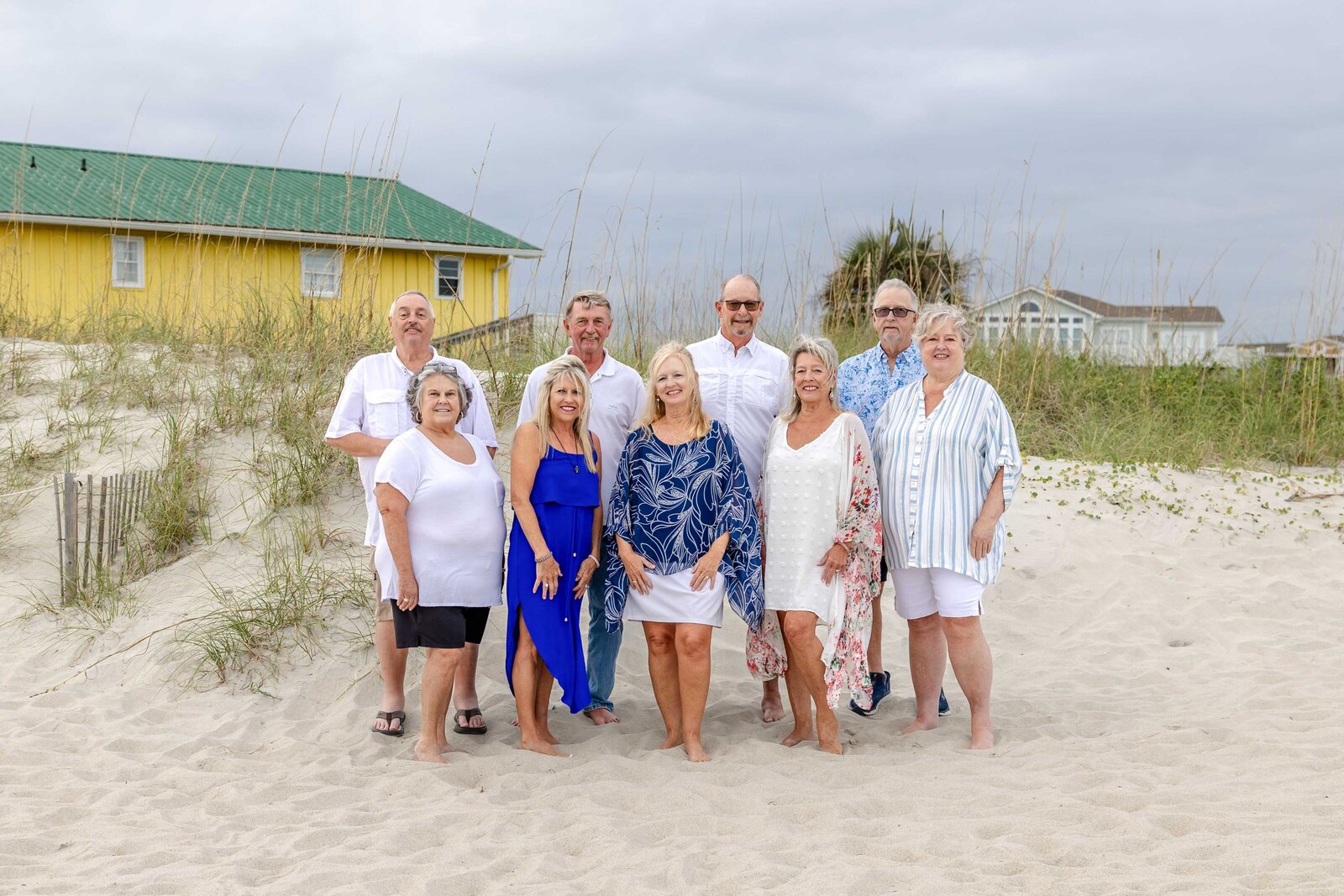A family poses on sandy dunes with a bright yellow beach house and sea oats in the background. Dressed in blue and white, they enjoy a relaxed coastal setting, perfect for artistic family photography in Wilmington, NC, highlighting beachside charm.