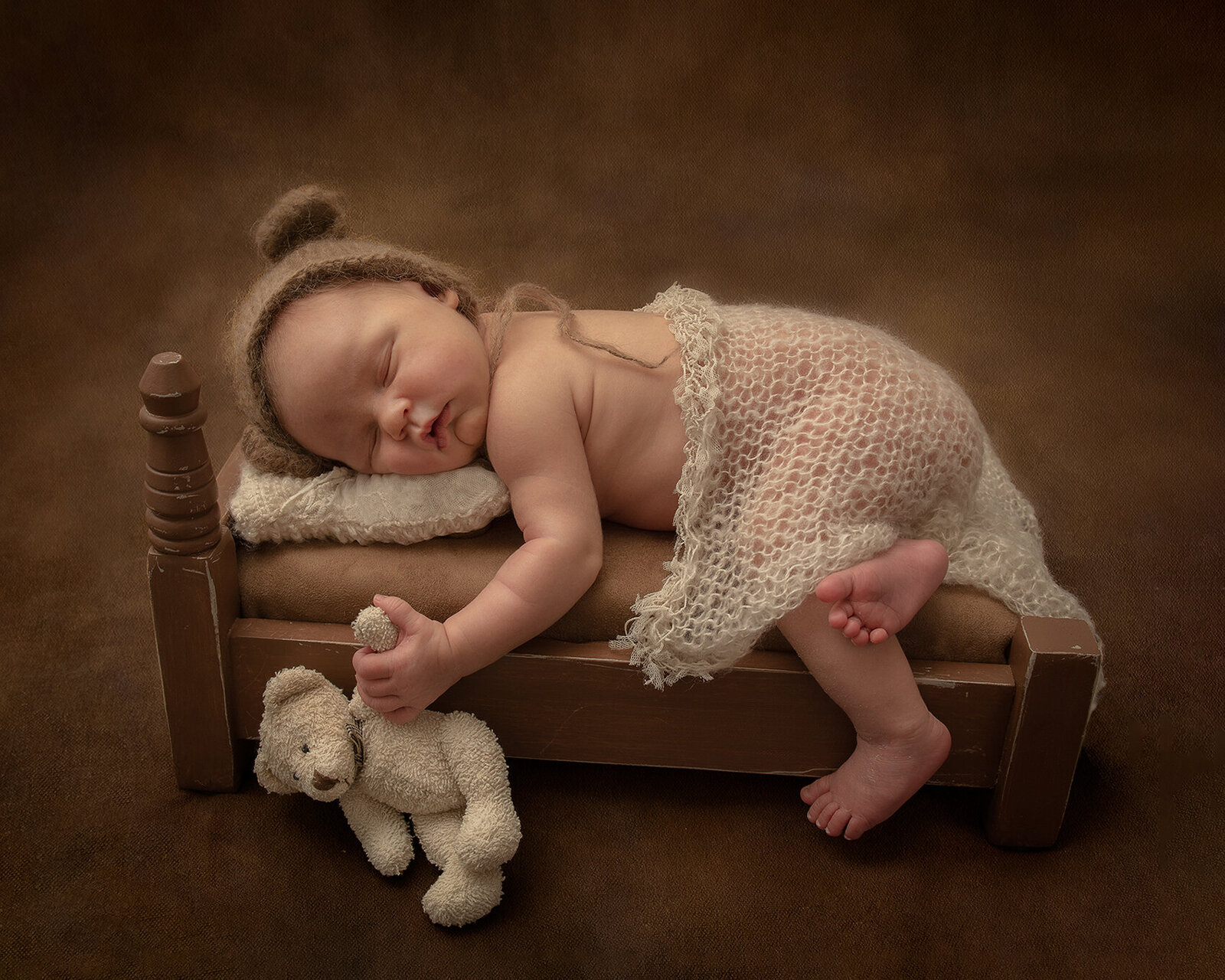 Baby girl on  a little bed, holding a stuffed bear and wearing a bear bonnet.
