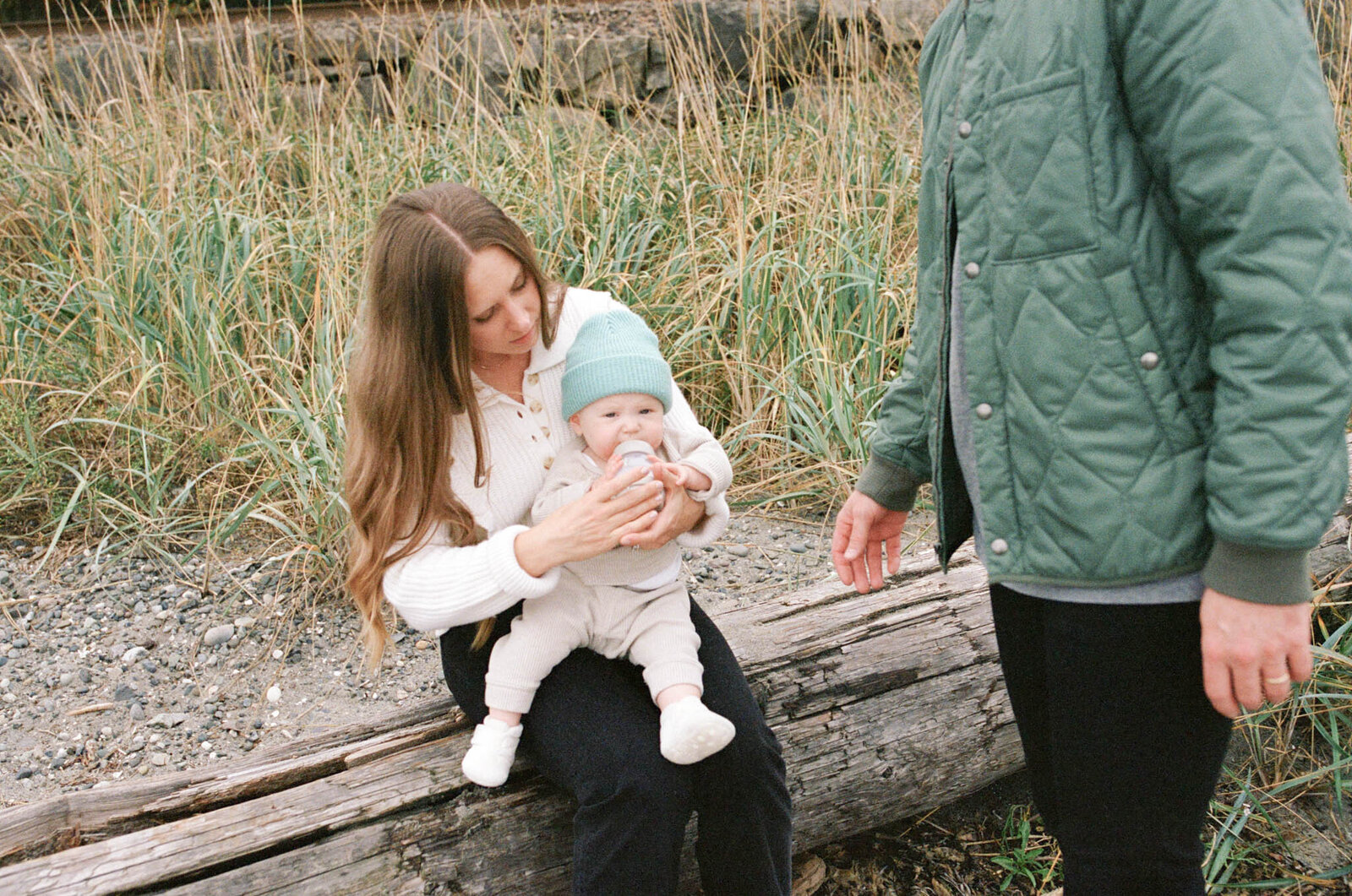 Mother giving baby girl a bottle while sitting on log at the beach on the Oregon Coast.