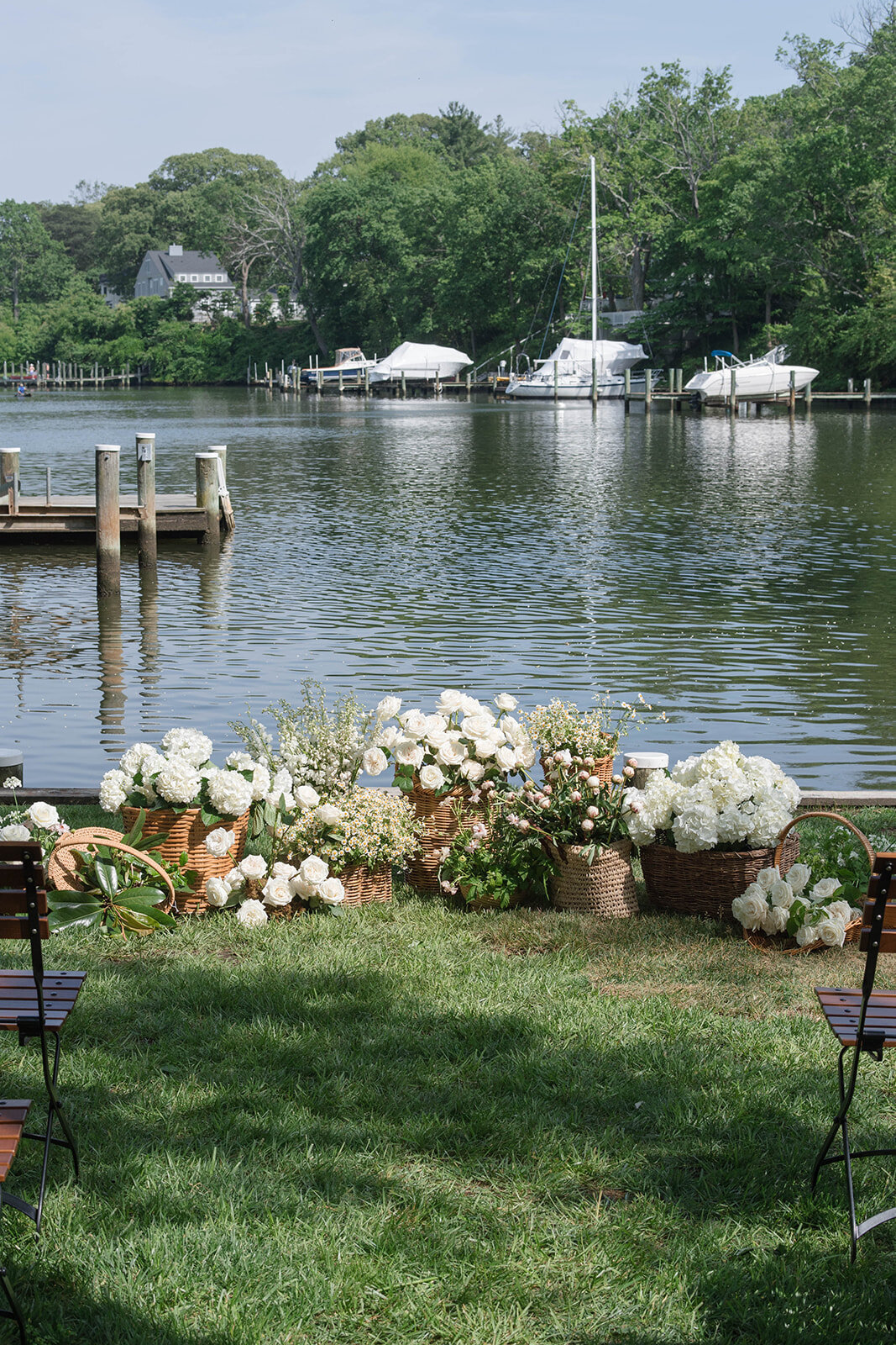 Ceremony display of assorted wicker baskets filled with white flowers including hydrangea, white garden roses, and tulips in front of a waterfront backdrop.