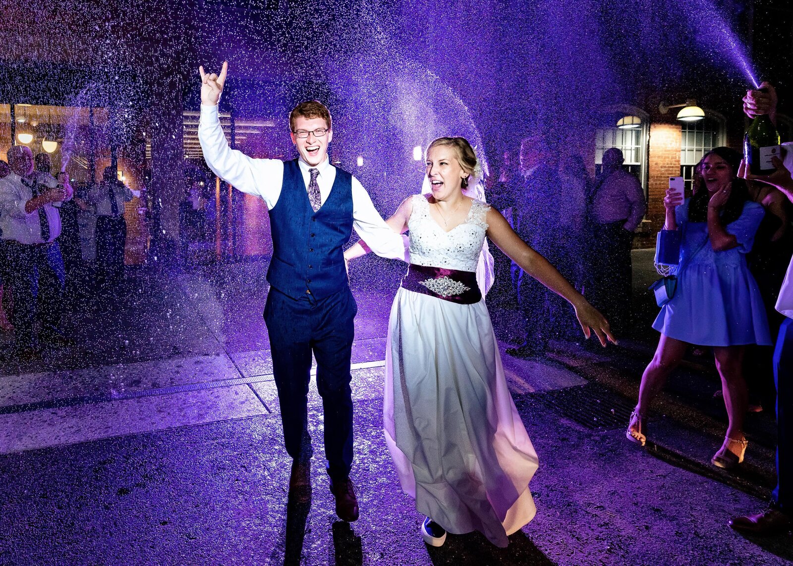newlyweds exit their wedding reception amidst champagne spray. they are backlit with purple lighting
