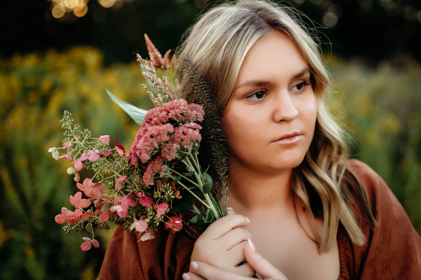 A person with long, blonde hair stands in a natural setting, holding a bouquet of pink and red flowers. The background is blurred greenery, creating a serene, outdoor atmosphere.