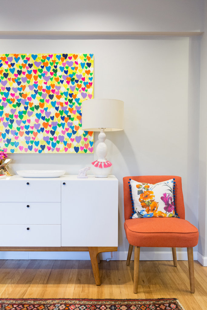 A modern white side table, colorful heart painting, and orange chair.