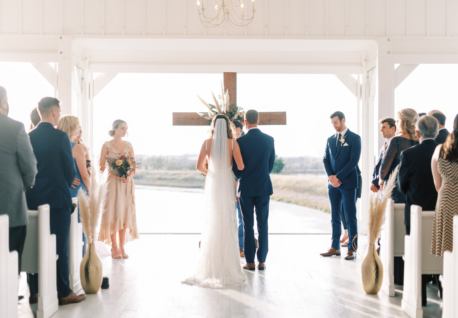 Portrait of bride and groom in a white wedding gown and black tuxedo standing at the altar in front of a cross.