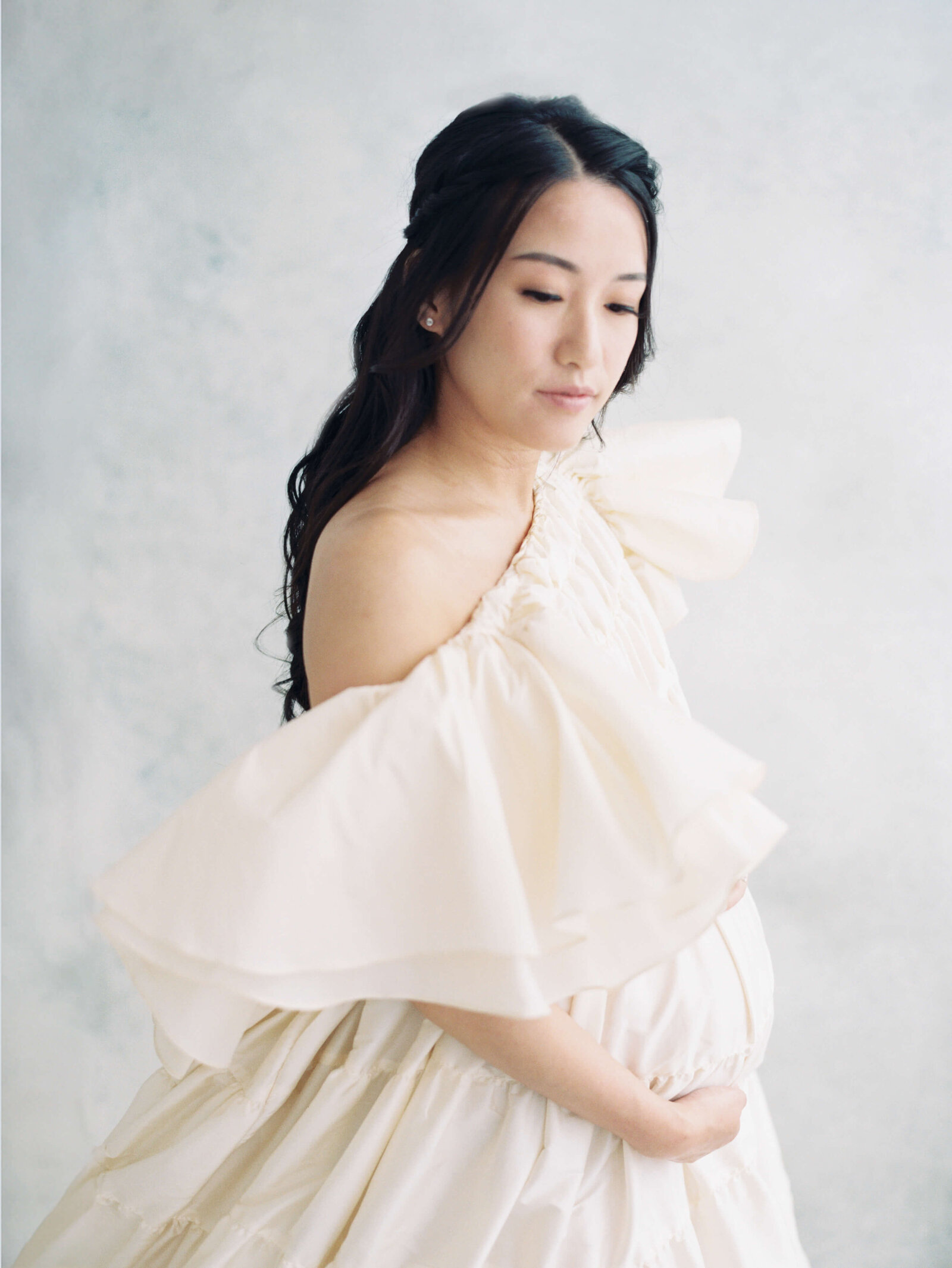 Expectant mother in a flowing gown, smiling warmly as she poses against a neutral studio backdrop.