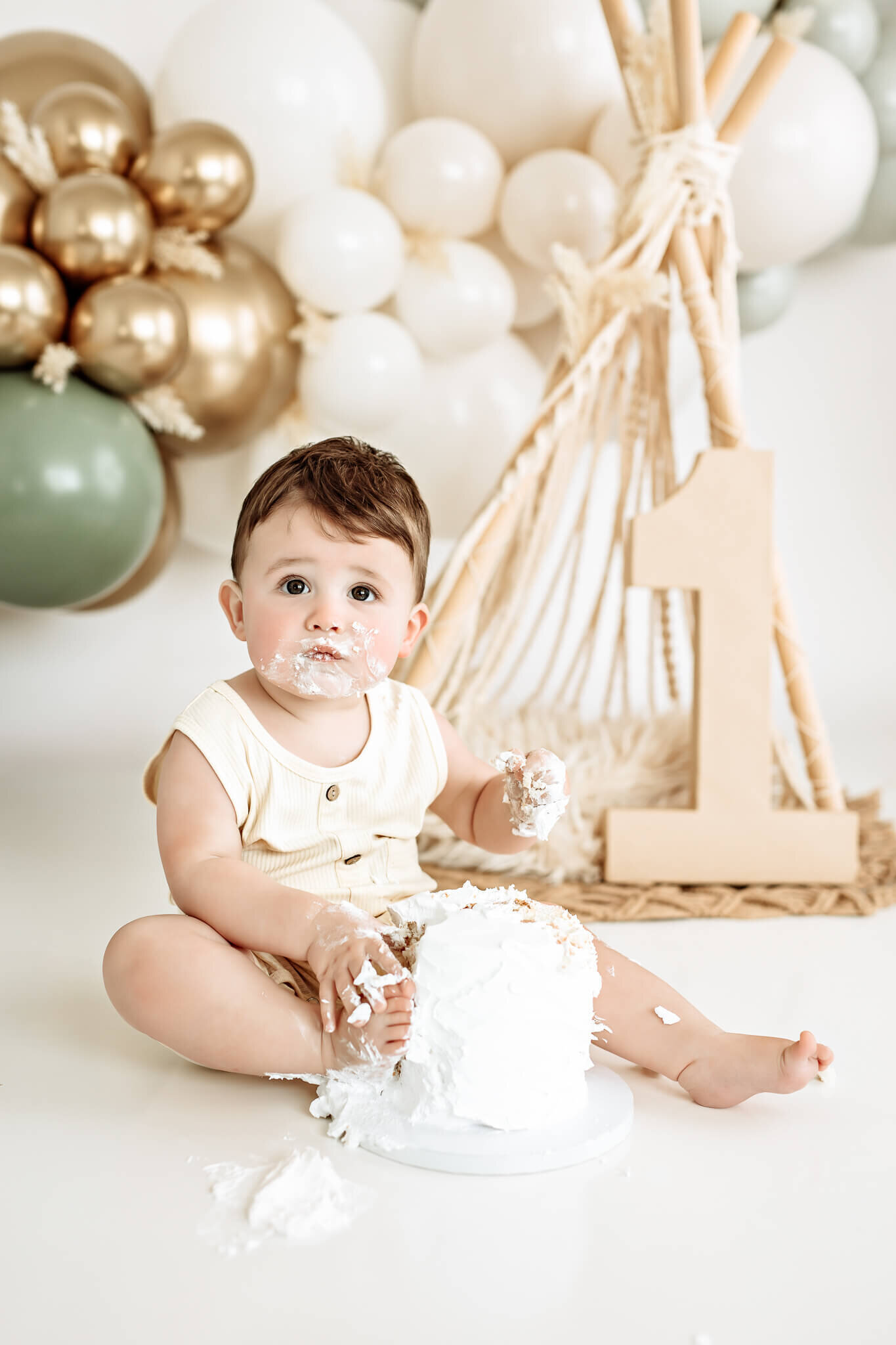 A baby sits on the floor with their hands in a large white frosted cake. They are wearing a light-colored outfit, and there is a cluster of balloons and a large number one decoration in the background.