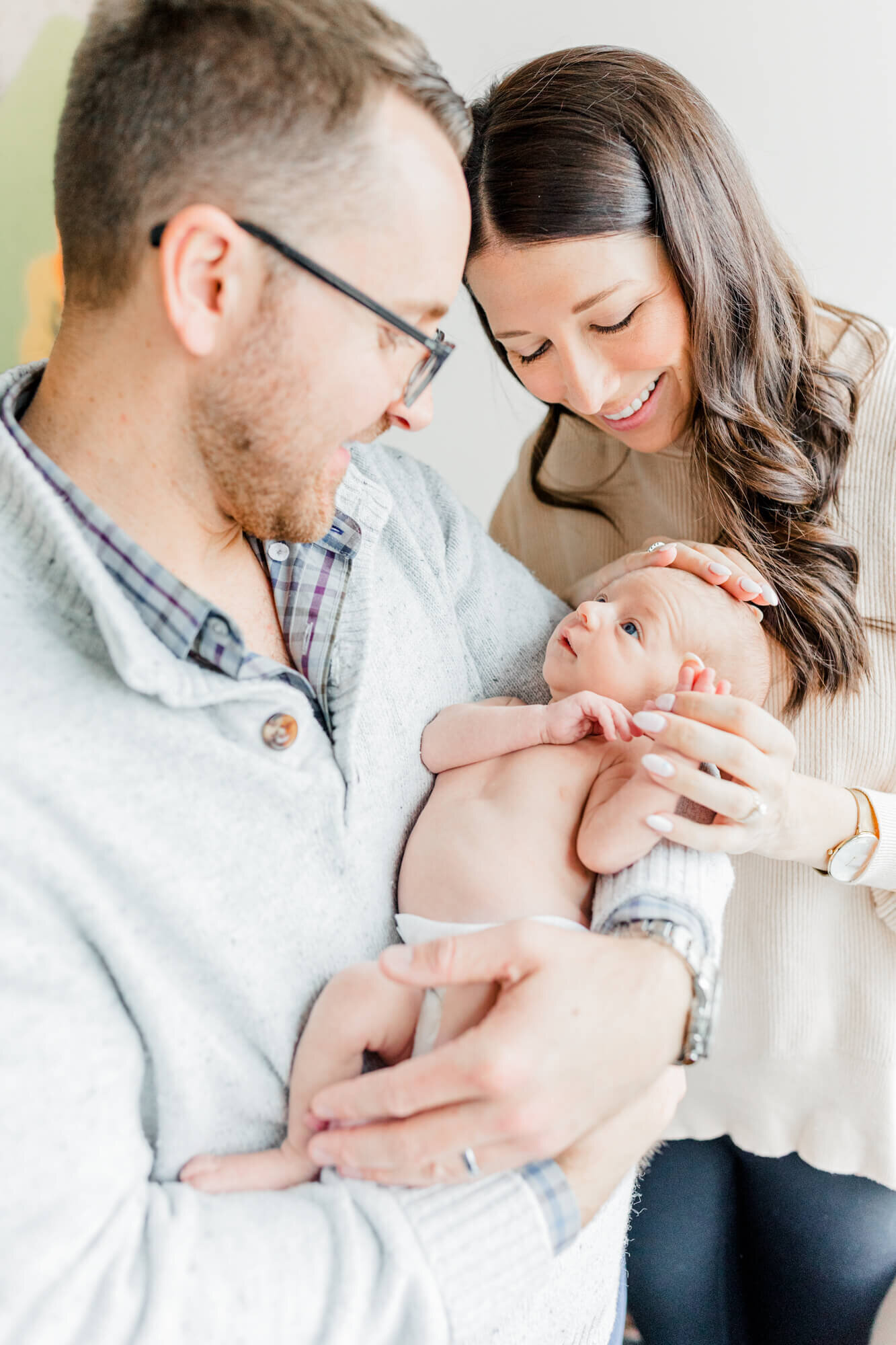 Newborn in his dad's arms looks curiously up at his dad while his smiling mom gently rubs his head