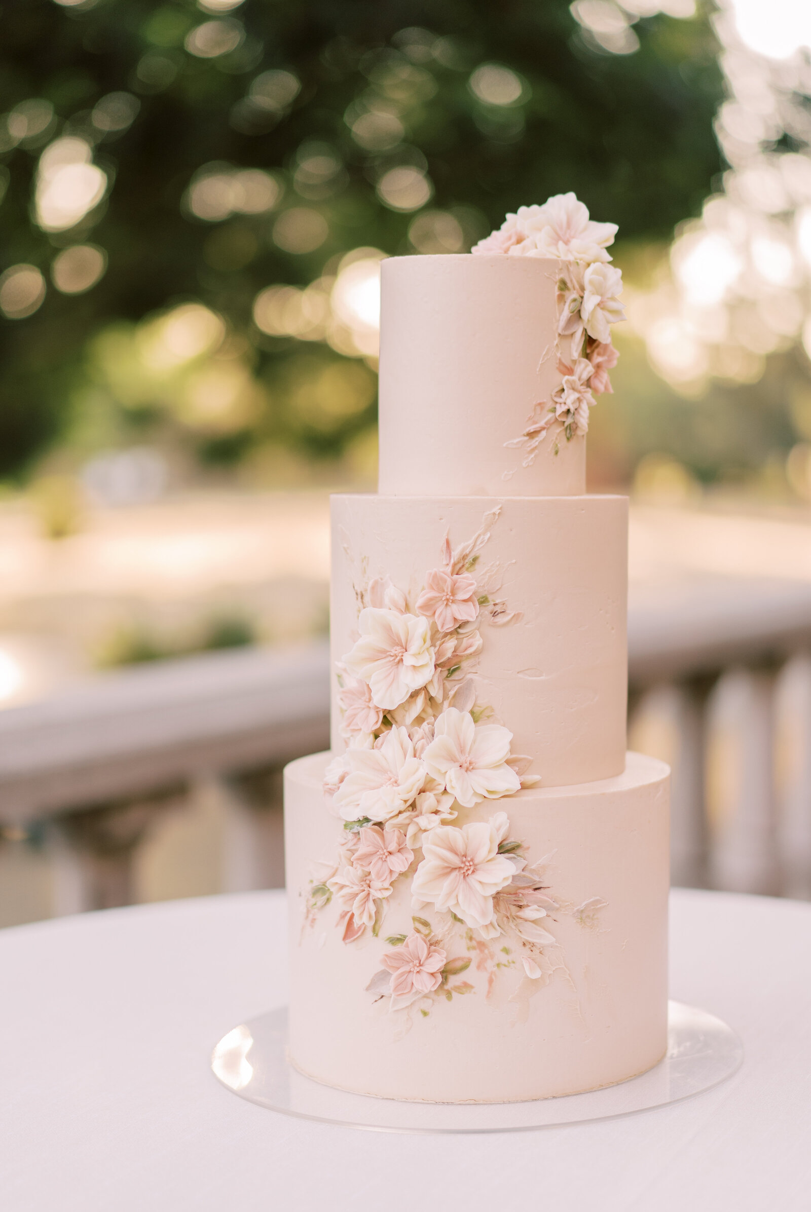 Portrait of a blush three-tiered wedding cake with flowers atop a round table outdoors.