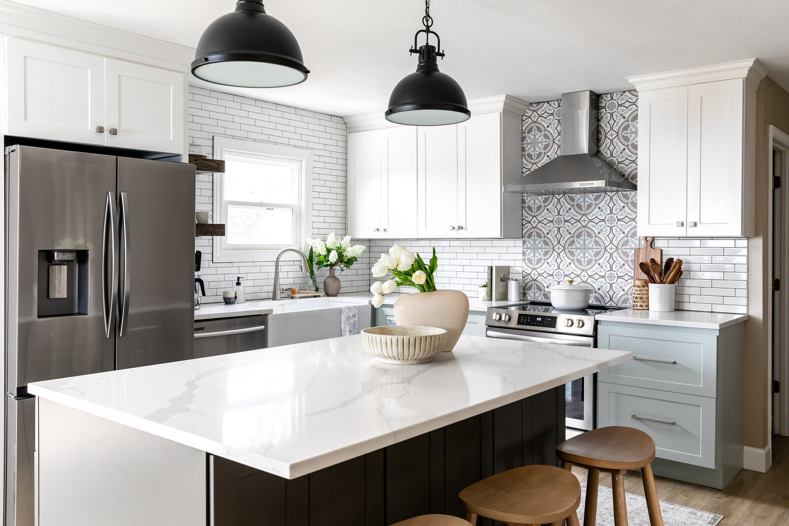 Light blue and white two-toned kitchen with subway tile and decorative tile above stove. Large island centered in room with two pendant lights. Large farmhouse sink below window.