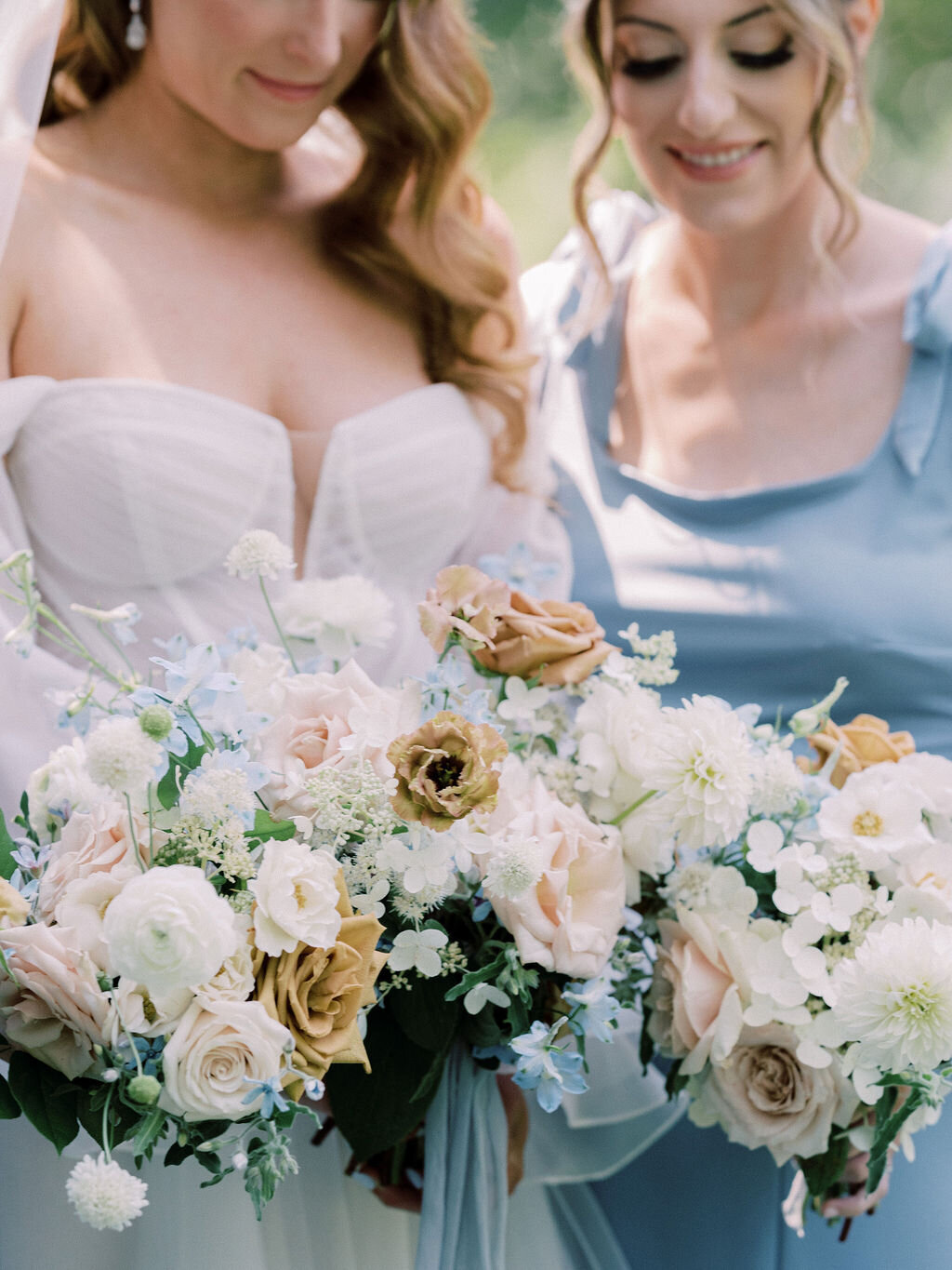Bride and bridesmaid holding bouquets of peach garden rose, white dahlia, toffee roses, white hydrangea, light blue delphinium and brown lisianthus and blue silk ribbon.