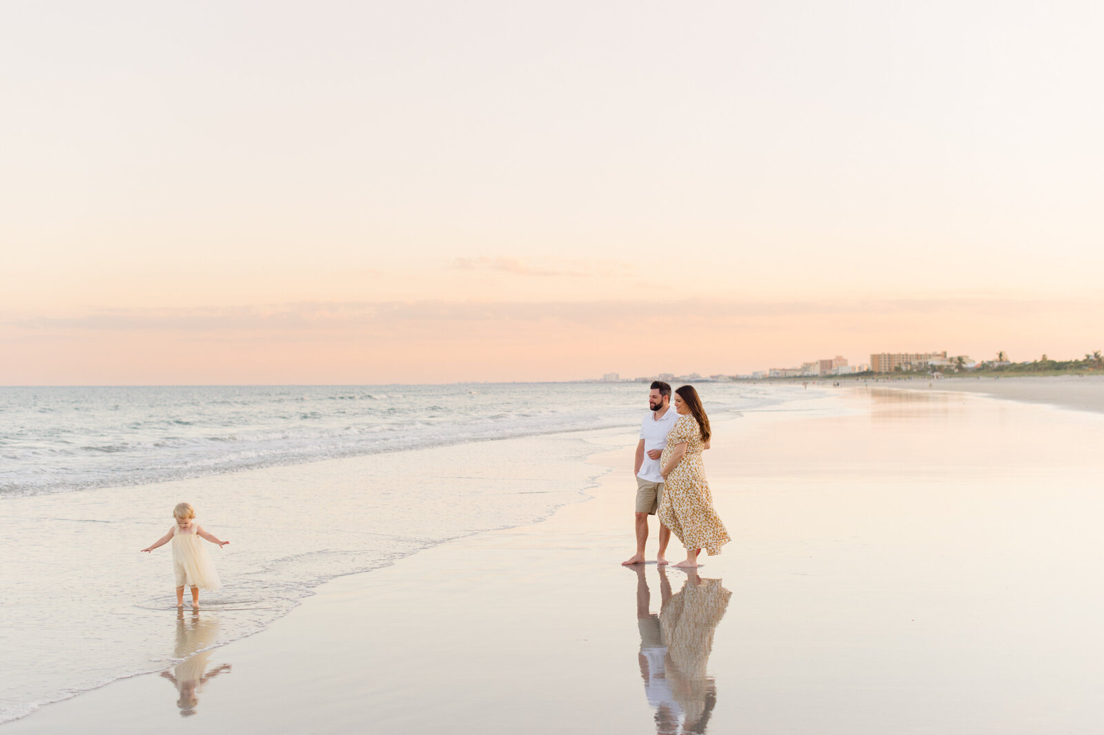 Pregnant couple watches their young daughter play in the ocean water at sunset in Cocoa Beach, Florida!