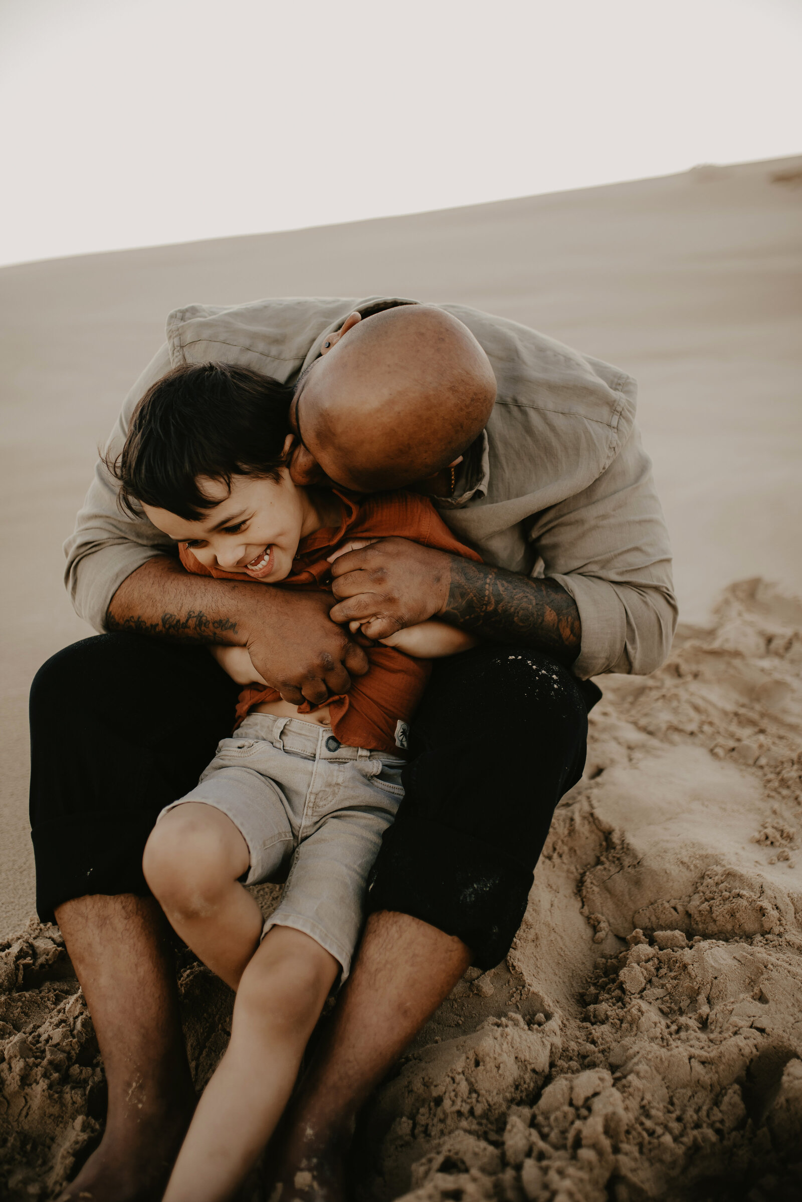 A bald man hugs and playfully wrestles with a young boy on a sandy dune. Both are smiling and appear to be enjoying the moment.