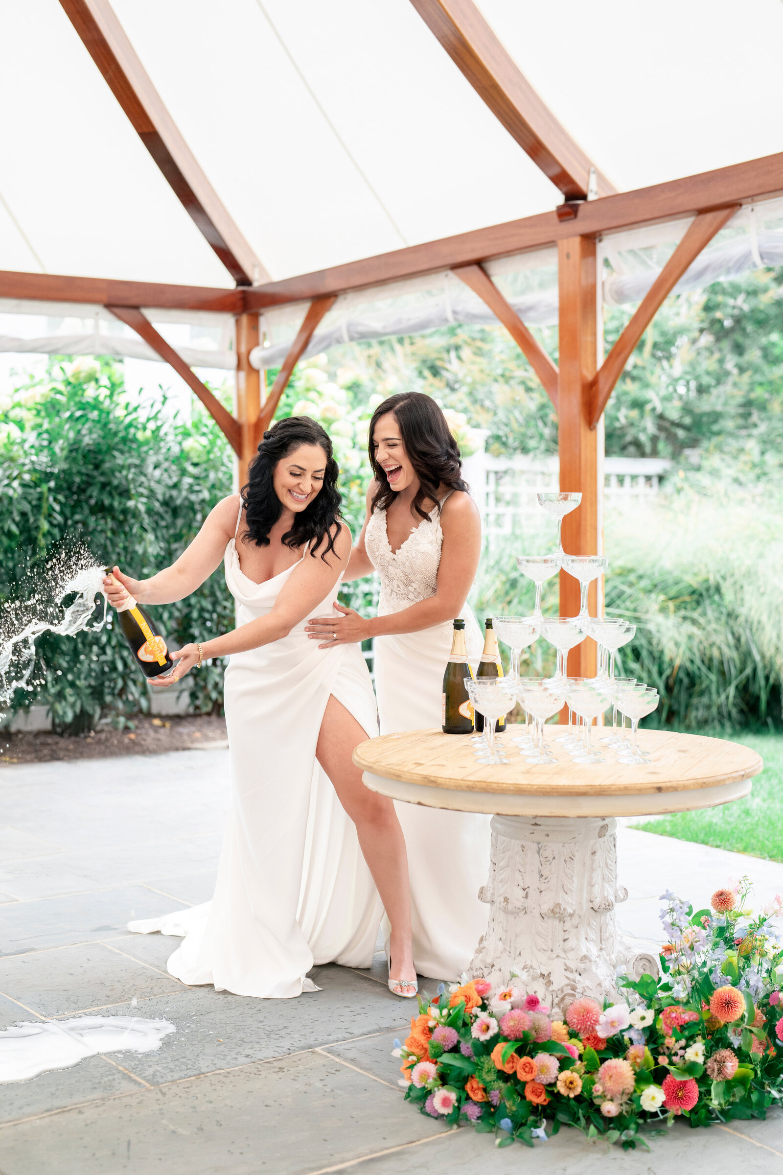 Two women in white dresses are joyfully pouring champagne into a tower of glasses on a round table adorned with colorful flowers. They are under a wooden canopy in a garden setting, with greenery in the background.