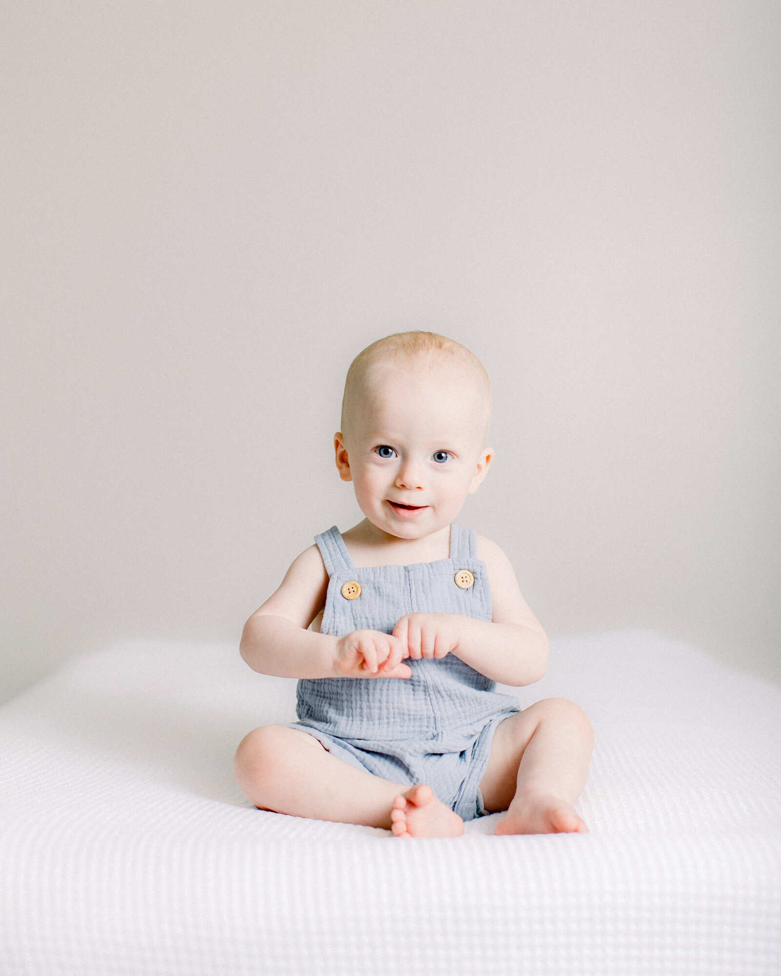 Happy baby sitting up and clasping his hands on white bedding by NH newborn photographer Fieldstone Studio.