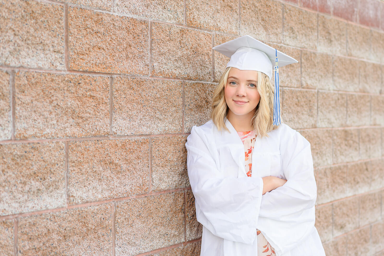 A high school senior girl wearing a white graduation cap and gown stands against a brick wall at North Lake Park in Lehi