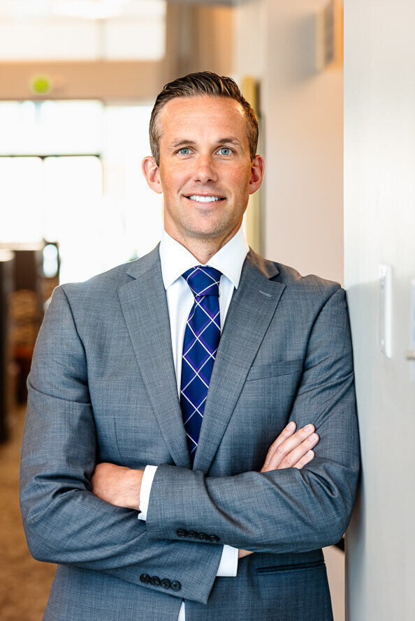 Man with gray jacket and blue tie folds arms and leans against wall.