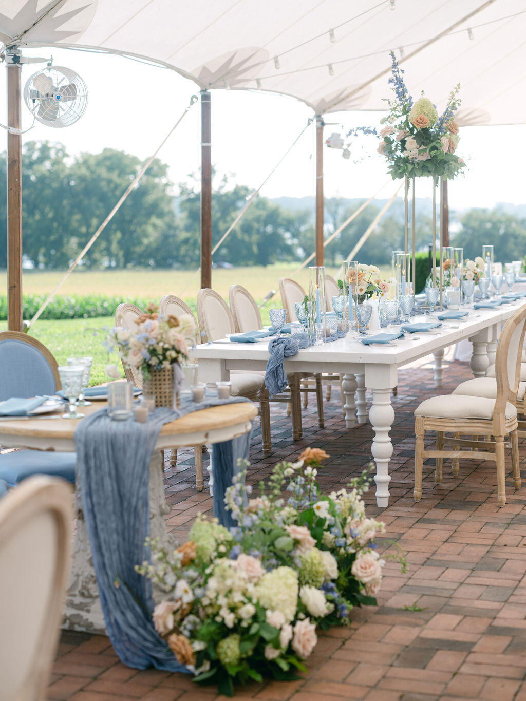 Sweetheart table besides the head table with florals to include green hydrangea, blue delphinium, toffee roses, blush garden roses, and white cosmos inside tented reception at Brittland Estate.