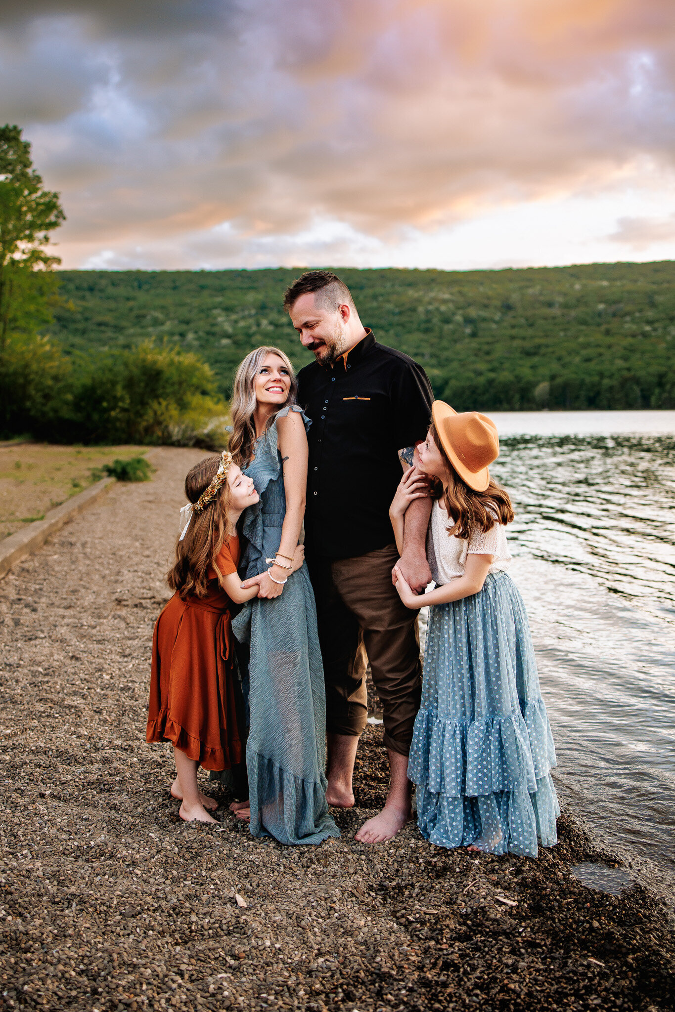 Along a beach with a gorgeous sunset in the background,  a mother and father gaze at each other, their two daughters snuggled on either side of them.