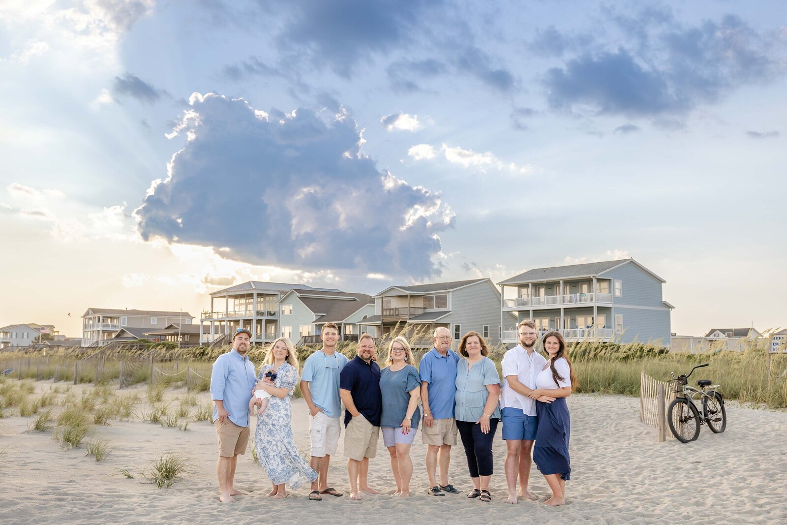 A family stands on the beach in front of beachfront homes, under a dramatic cloud formation. Dressed in blue and white, they enjoy the coastal scenery. Perfect for professional family photography in NC, highlighting beachside gatherings and stunning skies.