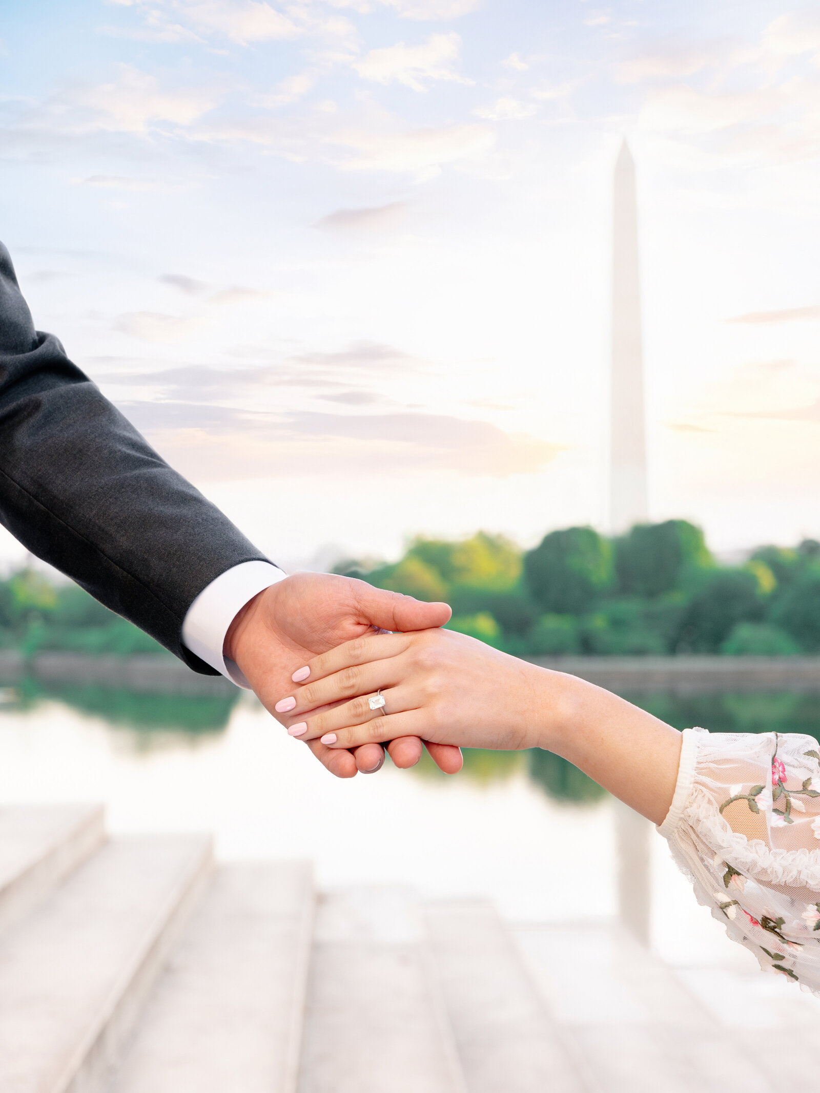 A couple holds hands, with the woman wearing an engagement ring. They stand on steps near a reflective body of water. The background shows a tall monument and a partly cloudy sky.