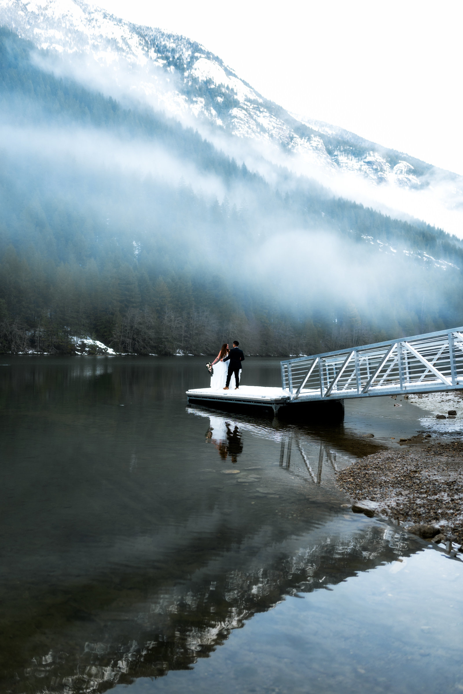 Couple on a dock on a lake, holding each other with snow covered mountain in background.
