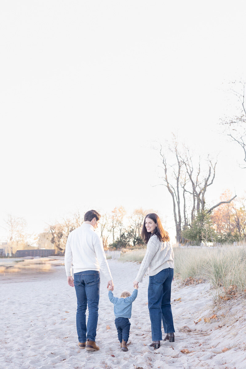 Young family walking hand-in-hand with their toddler on a quiet beach at sunset in Westchester County, New York. Captured by Nicole, a Connecticut and Westchester-based family photographer specializing in natural, relaxed moments.