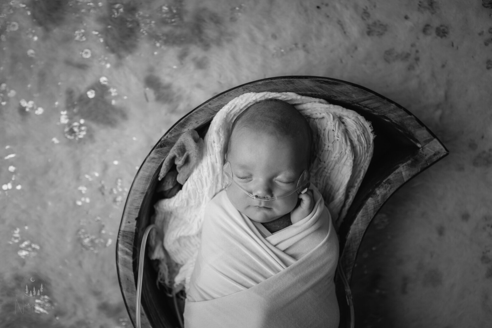 a black and white image of a newborn boy with oxygen use, in a wooden bowl with white wraps, on a sparkly cowhide style rug