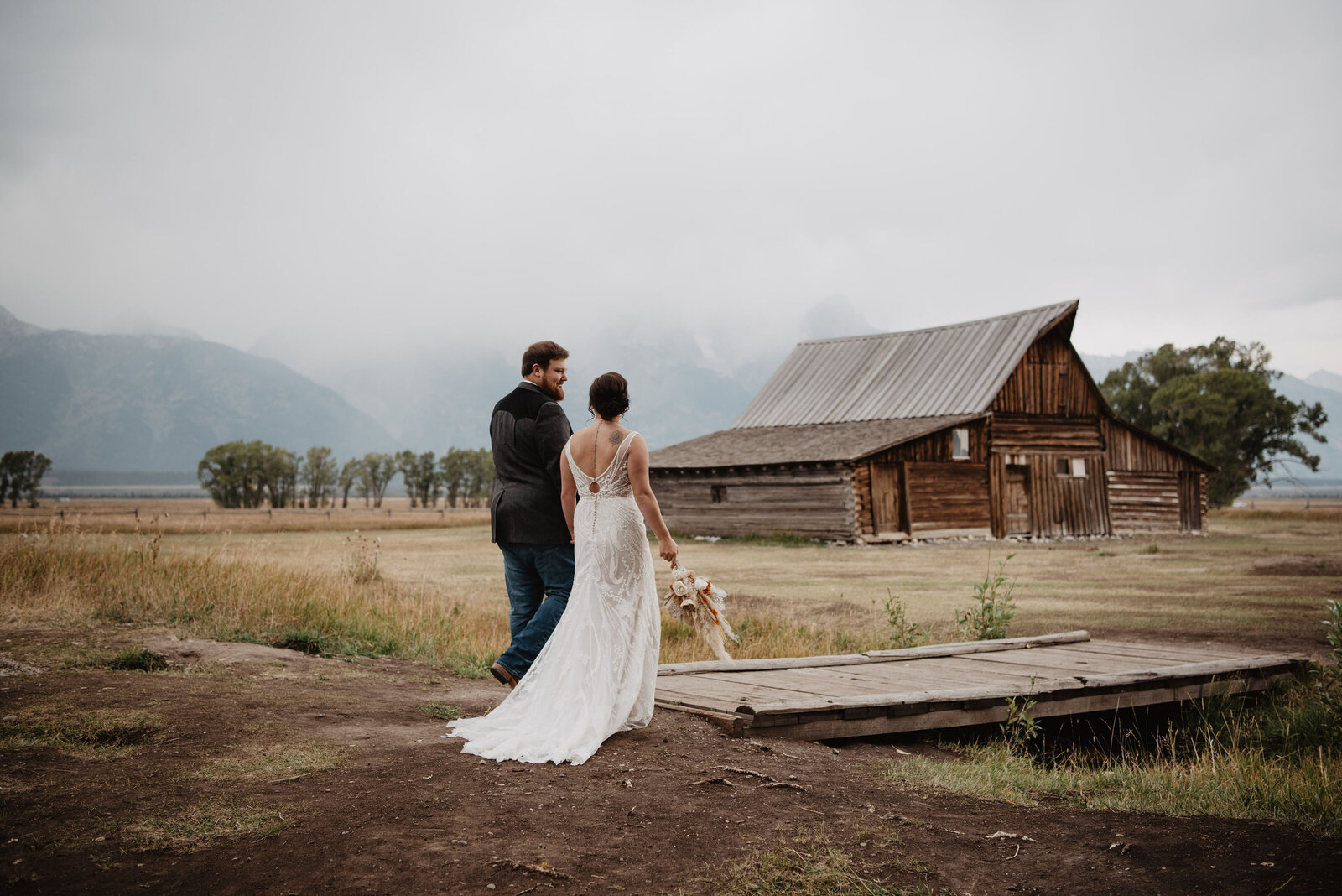 Mormon row in grand tetons in the fall with bride and groom walking together over a small bride next to a large barn for their jackson hole wedding  pictures on a foggy day
