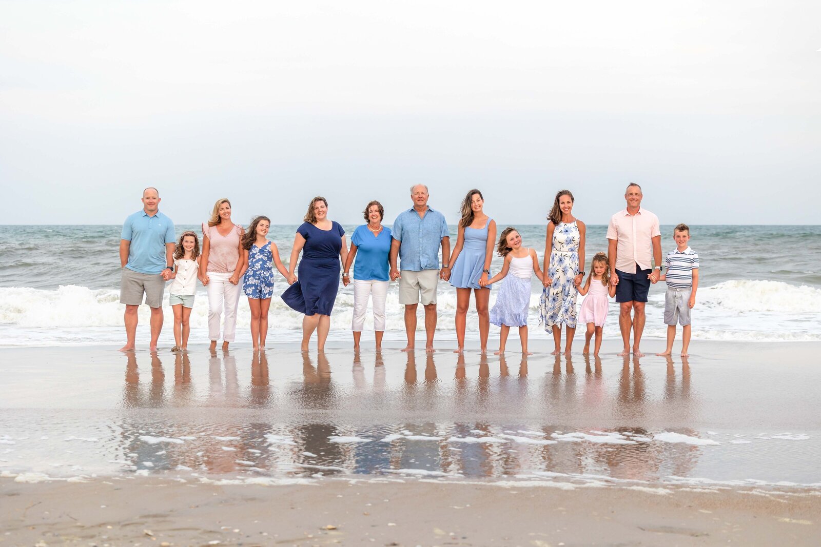  A large family stands together on the shoreline of Oak Island, reflecting in the wet sand. Dressed in blue and pastel colors, they capture the joy of a family reunion. Ideal for destination family photographer NC, showcasing vibrant beachside gatherings.