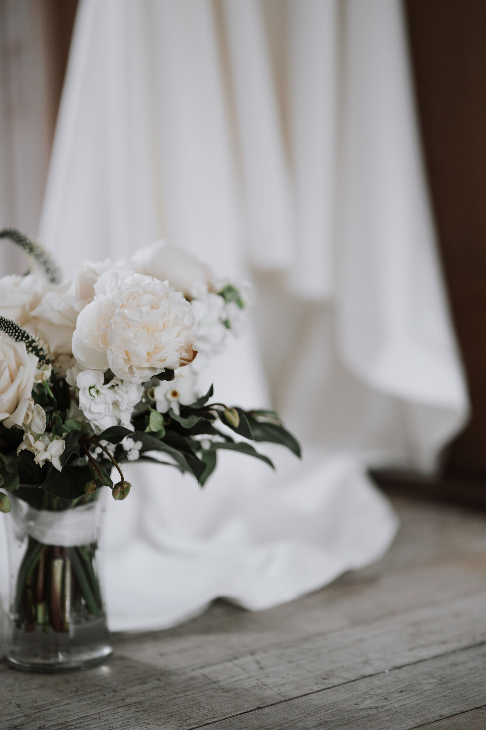 in the foreground the bridal bouquet shows off white roses wrapped with ribbon insode a vase in the background the  white wedding dress flows captured by Idaho Falls Wedding Photographer