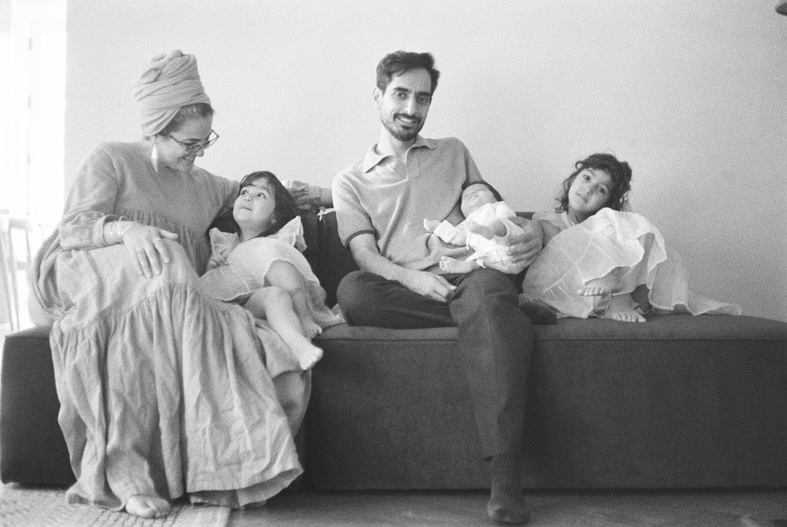 A cozy black-and-white family portrait of parents sitting on a couch with their three young children, capturing a moment of togetherness and warmth.