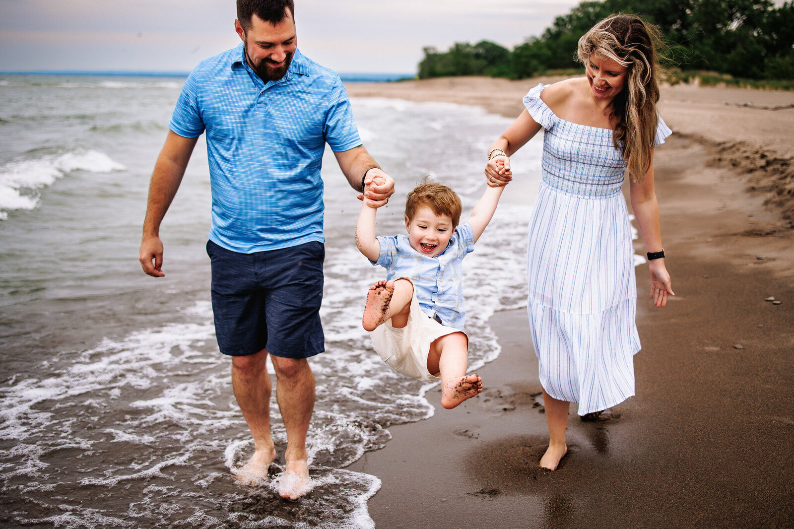 A mother and father walking along the beach swing their giggling toddler forward as a wave crashes amongst their feet 