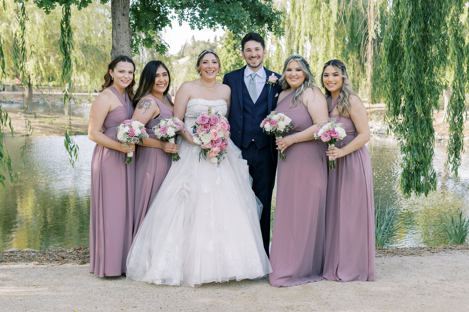 bride and groom posing with the bridesmaids in front of a lake surrounded by weeping willows with bridesmaids in mauve bridesmaids dresses captured by sacramento wedding photographer