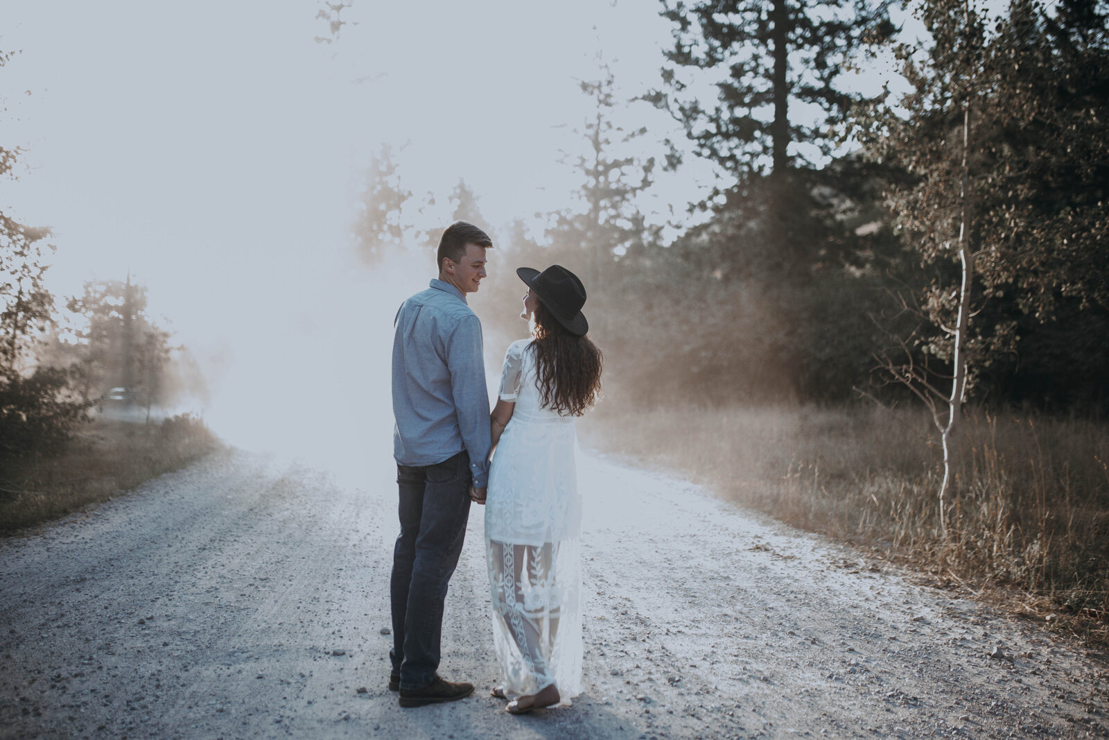 man and woman holding hands on a dirt path together while walking away from the camera into the fog on the woods
