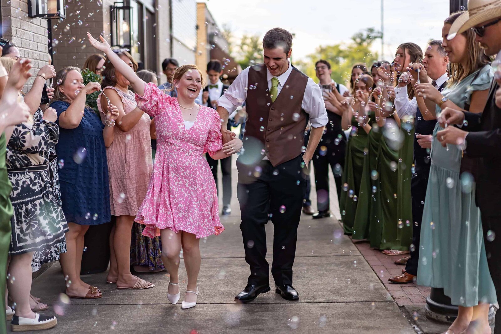 A couple waves joyfully in the center of a cheering crowd outdoors, surrounded by people blowing bubbles and clapping, dressed in semi-formal attire on a bright day in Ruston, Louisiana.
