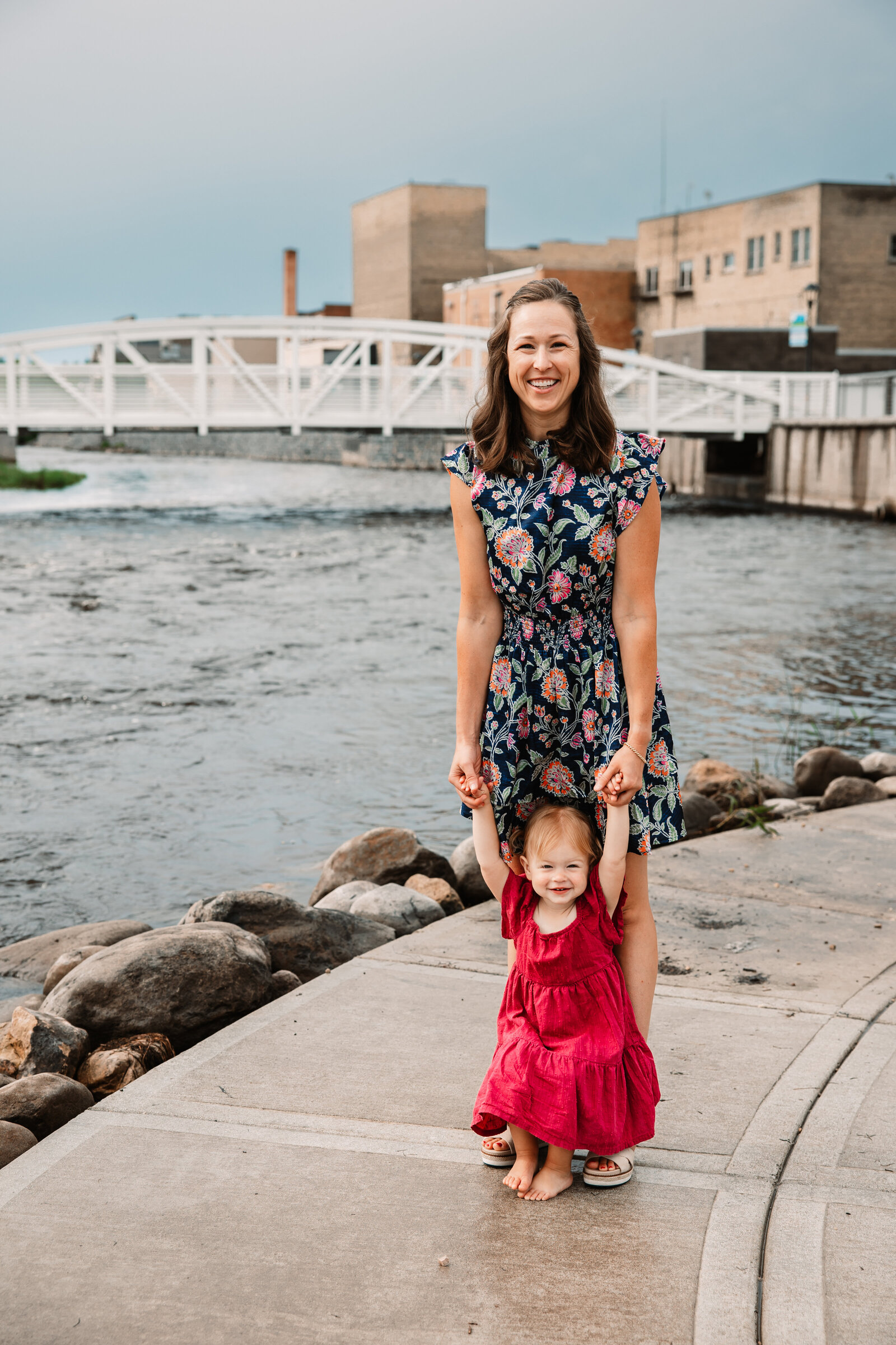 A toddler girl in a red dress walks on a lakeside sidewalk with help from mom