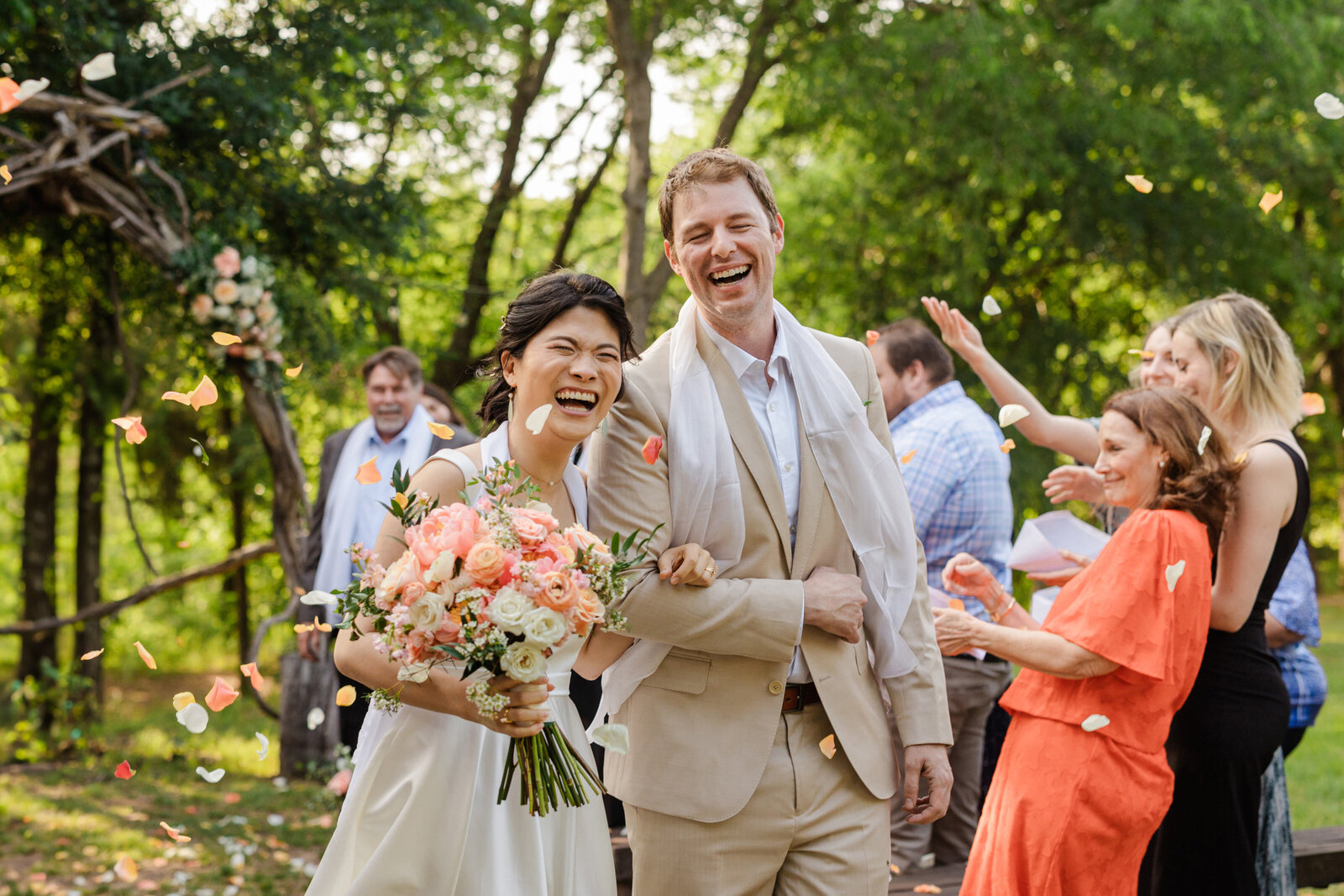 DFW Wedding Photographer; Asian bride and Caucasian groom  in formal wedding attire joyfully laughing arm in arm as they recess down the aisle after their wedding ceremony while their guests shower them with rose pedals