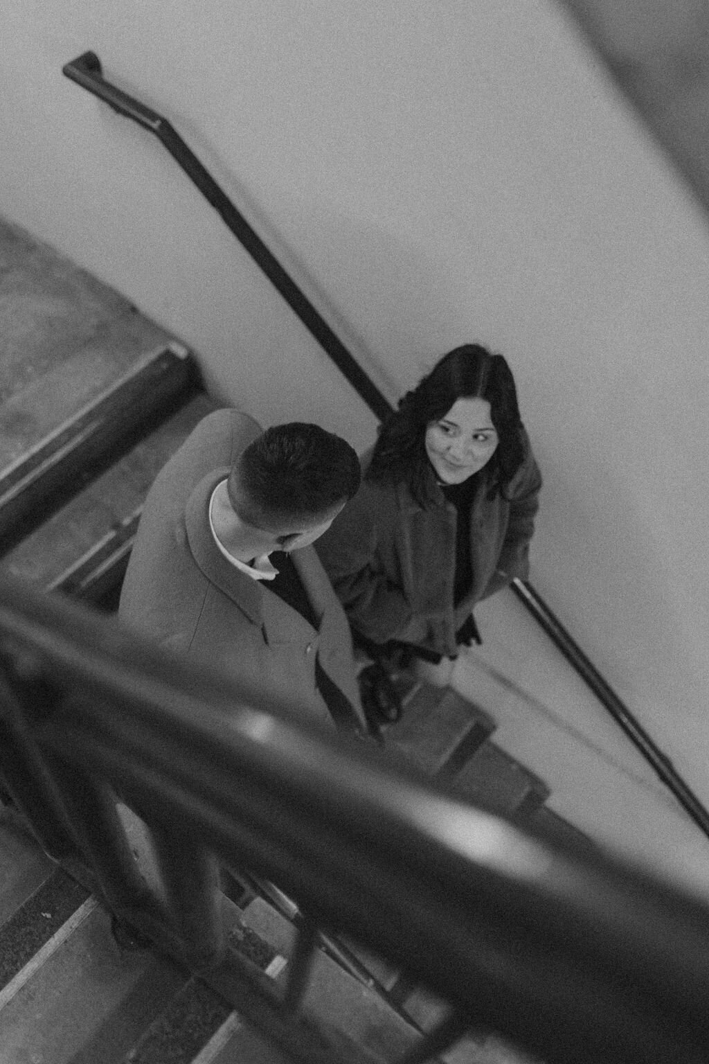 Black and white photo of couple in coats walking down industrial stairs looking sweetly at each other