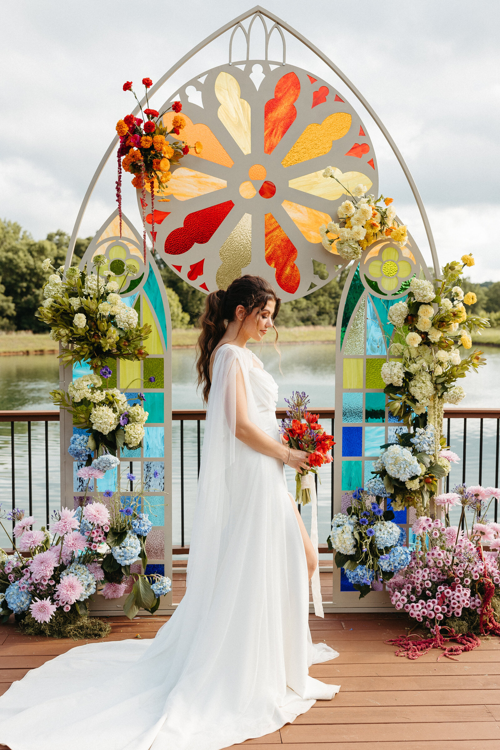 Bride standing in front of colorful stained-glass-inspired wedding altar with bold floral arrangements by the lake