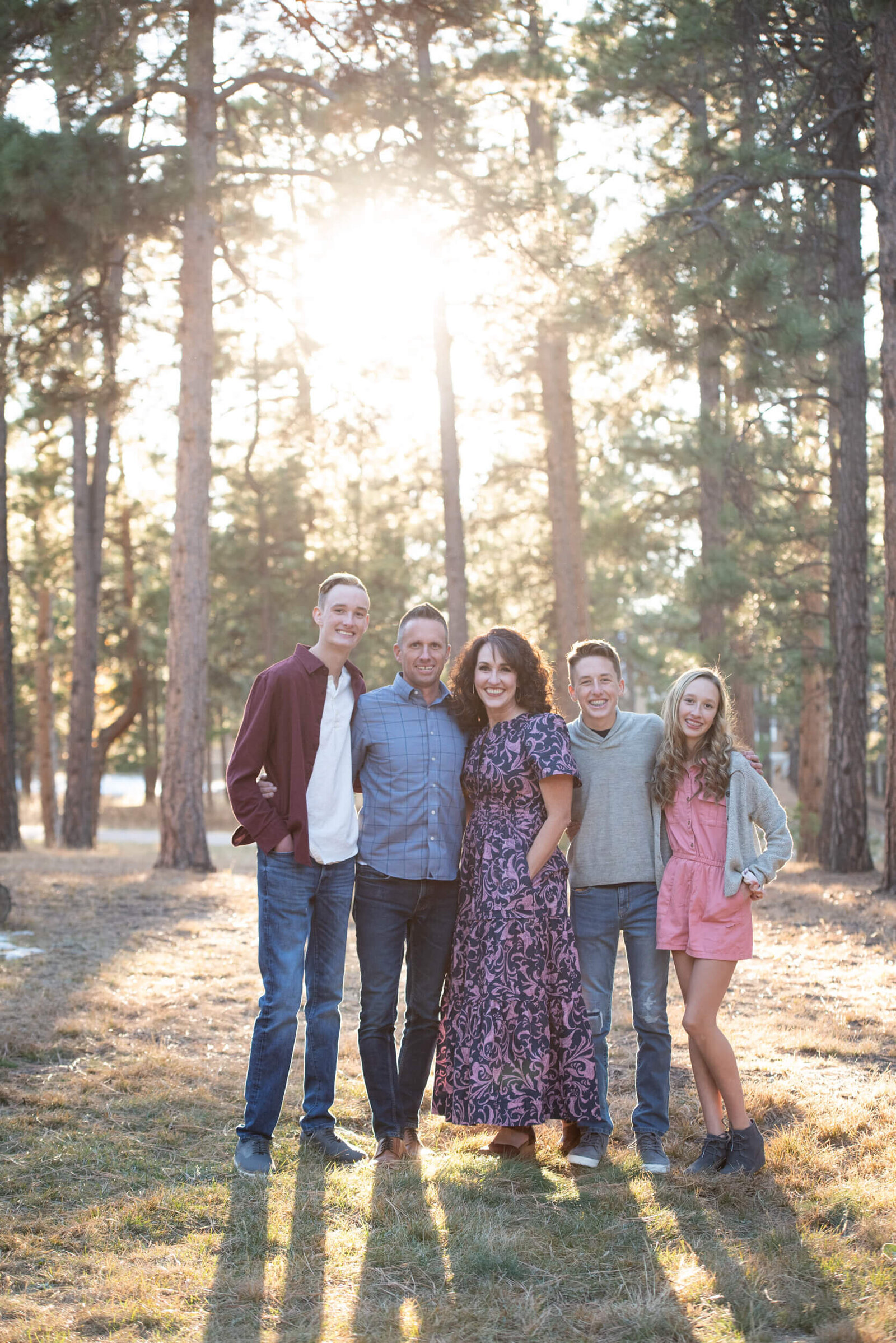 A smiling family of 5 stands with arms around each other in a park at sunset
