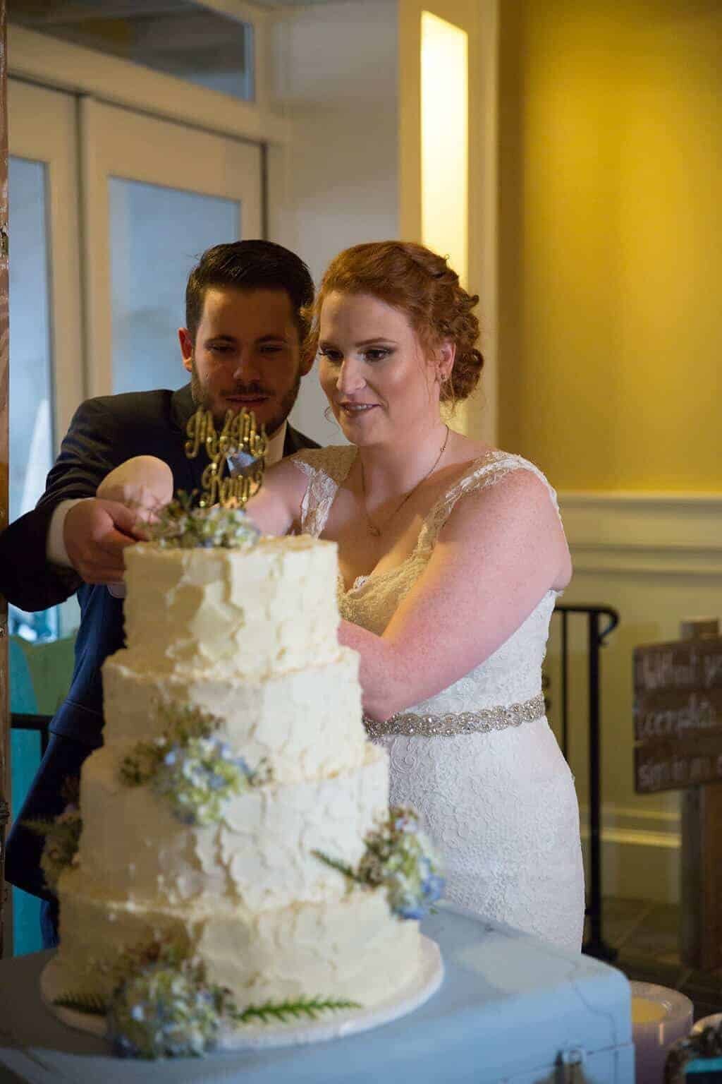 A bride and groom are cutting a three-tiered white wedding cake decorated with flowers. The bride, in a lace dress, and the groom, in a dark suit, are both holding the cake knife together.