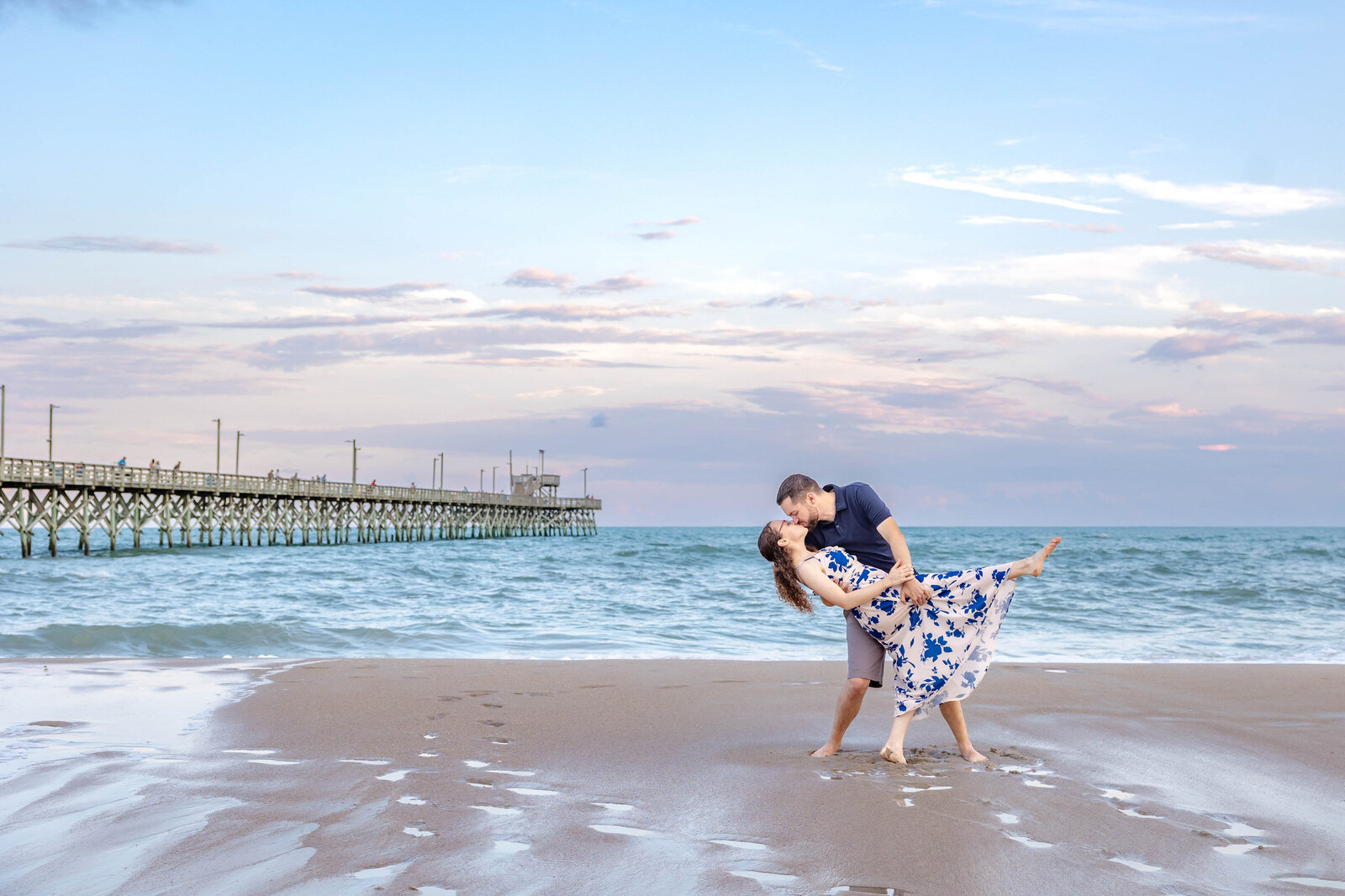 A couple shares a romantic dance pose on sandy North Topsail Beach, NC, with the man dipping the woman who wears a flowing floral dress. The serene ocean and a long pier under a pastel sky create a dreamy setting, showcasing the elegance of North Carolina engagement photography.  