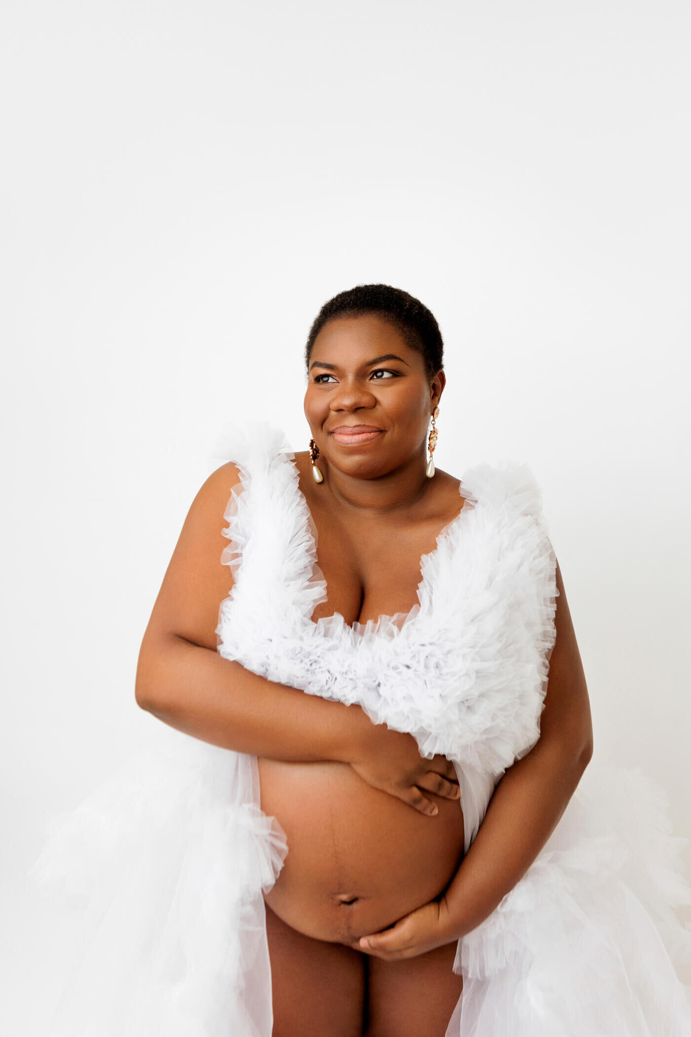 A pregnant woman stands against a white background, cradling her baby bump with both hands. She is wearing a luxurious, white tulle dress with ruffled details, and her expression is joyful and serene. She has short hair, elegant earrings, and a natural, radiant beauty that is highlighted by the soft lighting.