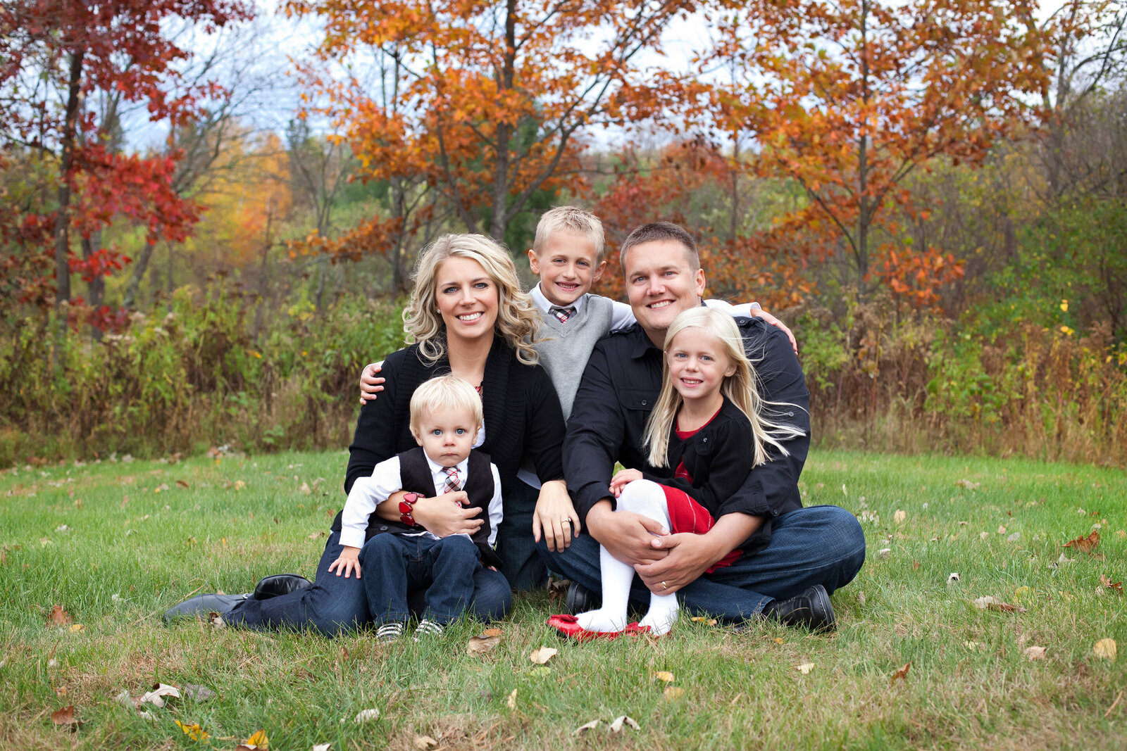 A young family of five sitting on the grass embracing with the fall colors behind them at Studio 64 Photography in Akeley, Minnesota.